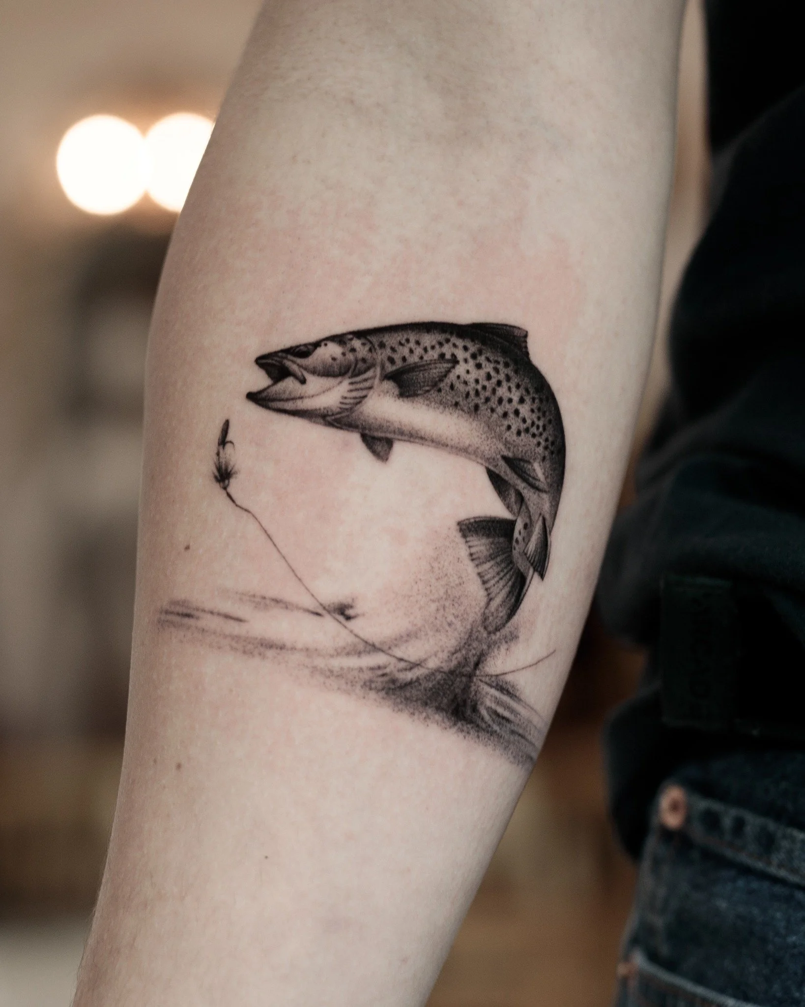 Black and grey forearm microrealism tattoo of a leaping trout approaching a fishing fly, with detailed spots, fins and flowing lines beneath.