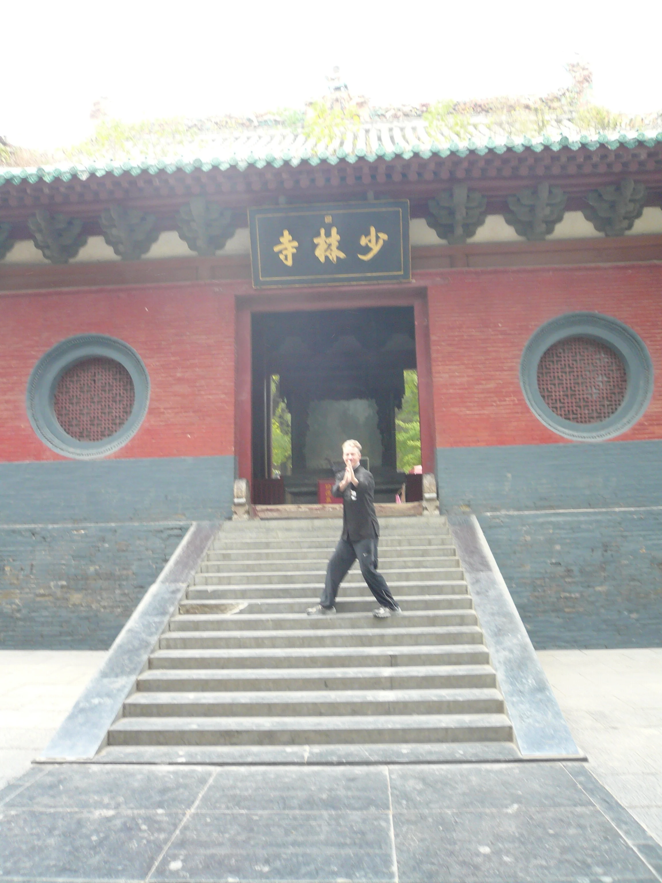 Sifu Kelly in a martial arts pose on stairs in front of a Chinese temple (Shaolin) with red walls, gray brick foundation, and black and gold sign with Chinese characters.