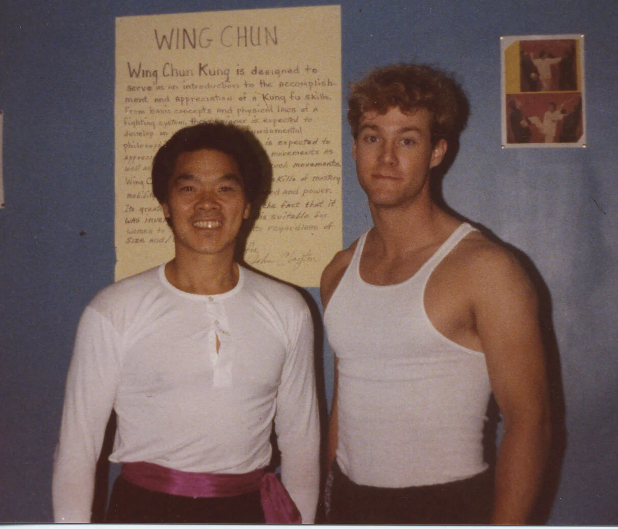 A young William Cheung (Grandmaster of Wing Chun Kung Fu) and a young Sifu Kelly standing side by side in front of a blue wall with a poster about Wing Chun martial art.