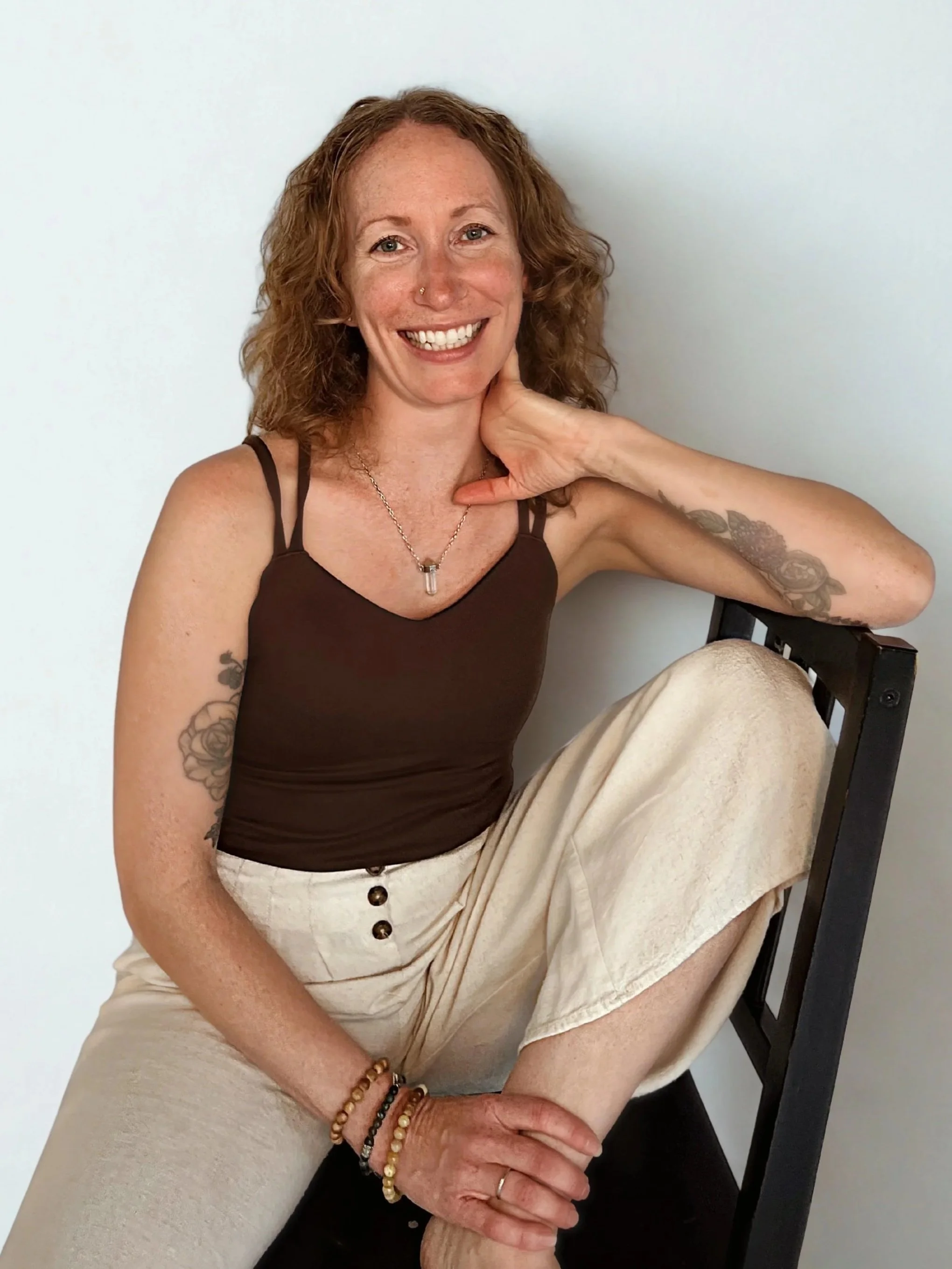 Portrait of a red-haired woman smiling, wearing a sleeveless brown top, black pants, and a watch, sitting against a plain white background.