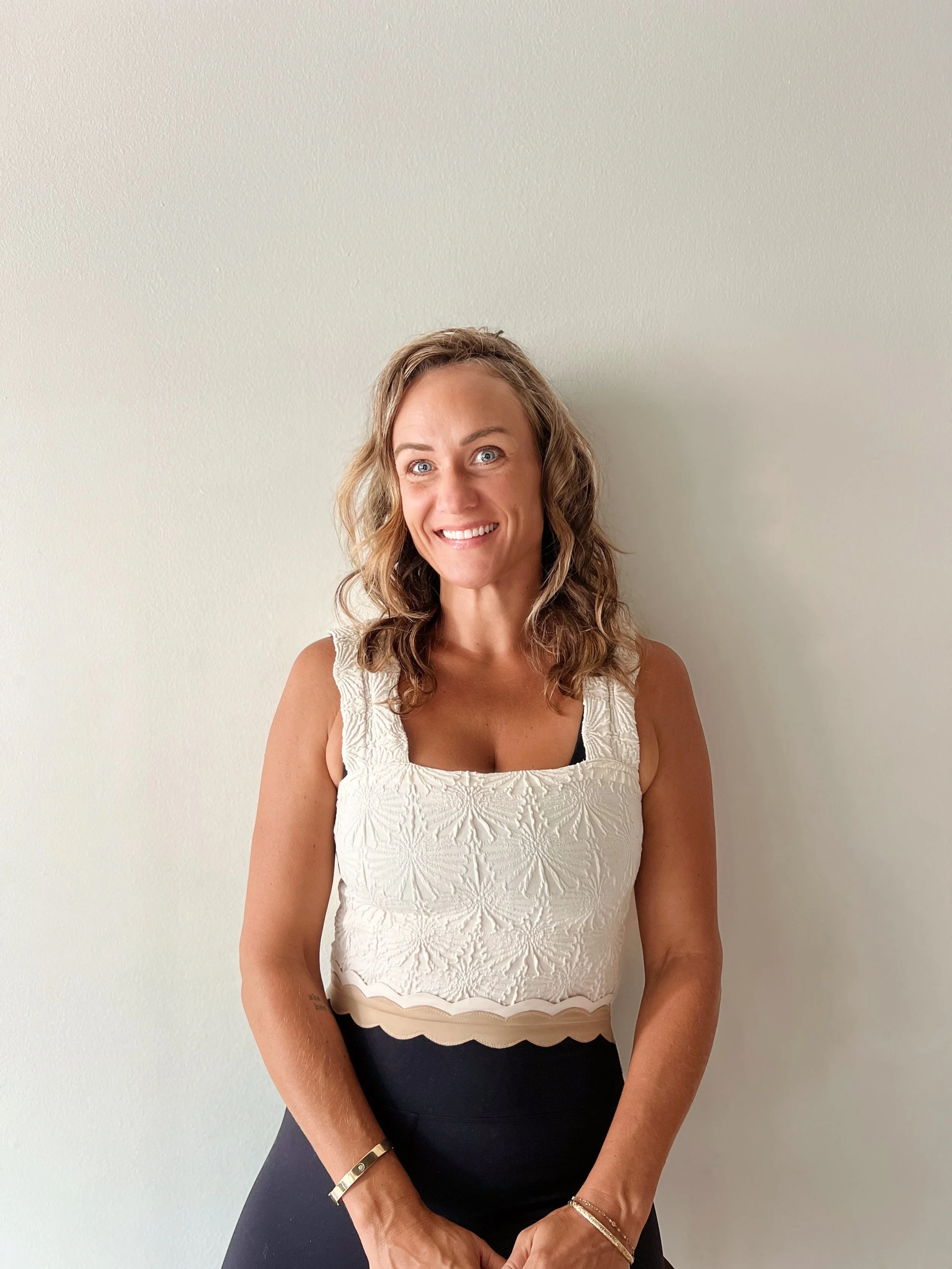 A woman with curly brown hair, wearing a white T-shirt and black tie-dye pants, smiling while sitting against a plain white wall.