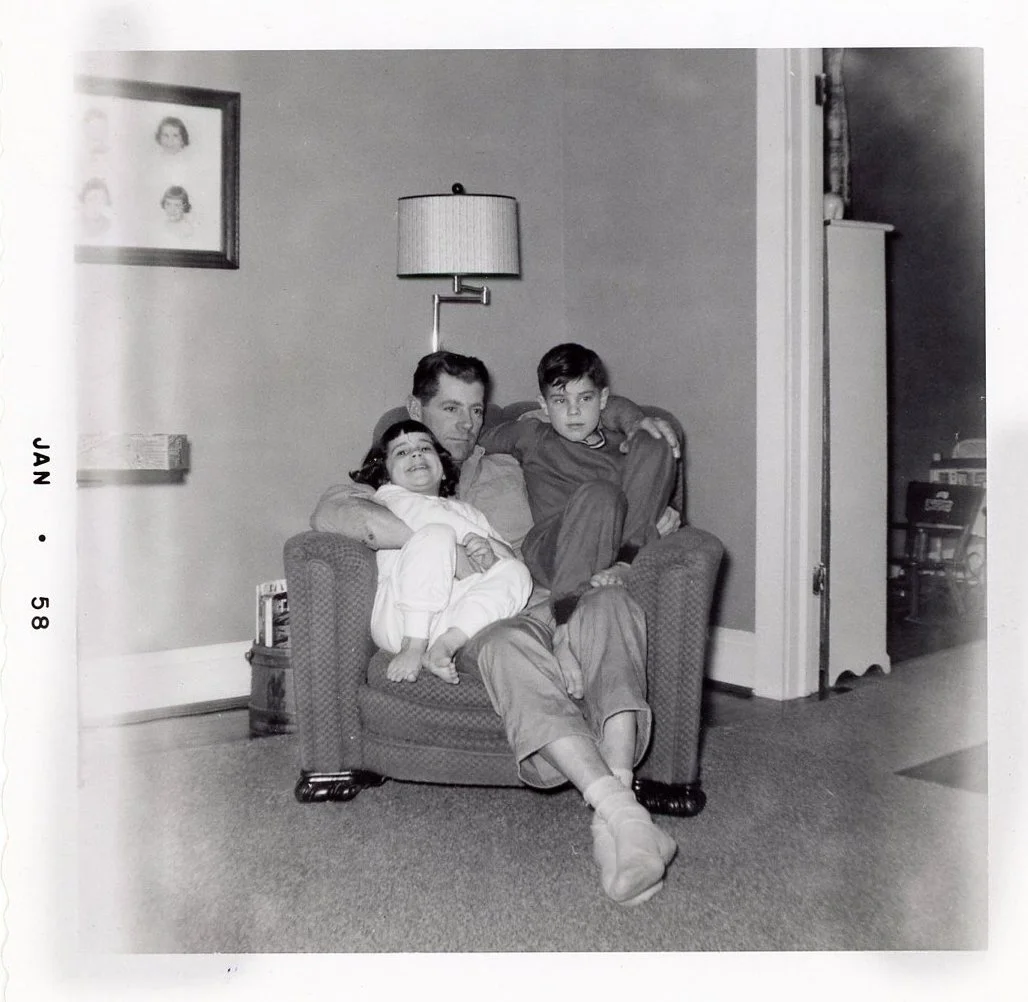 A black and white photo of a man sitting on a small armchair with two children, a girl and a boy, sitting on his lap, in a living room. The girl is smiling and the boy looks serious. There is a wall with a framed picture and a lamp behind them, and a partially open door to another room.