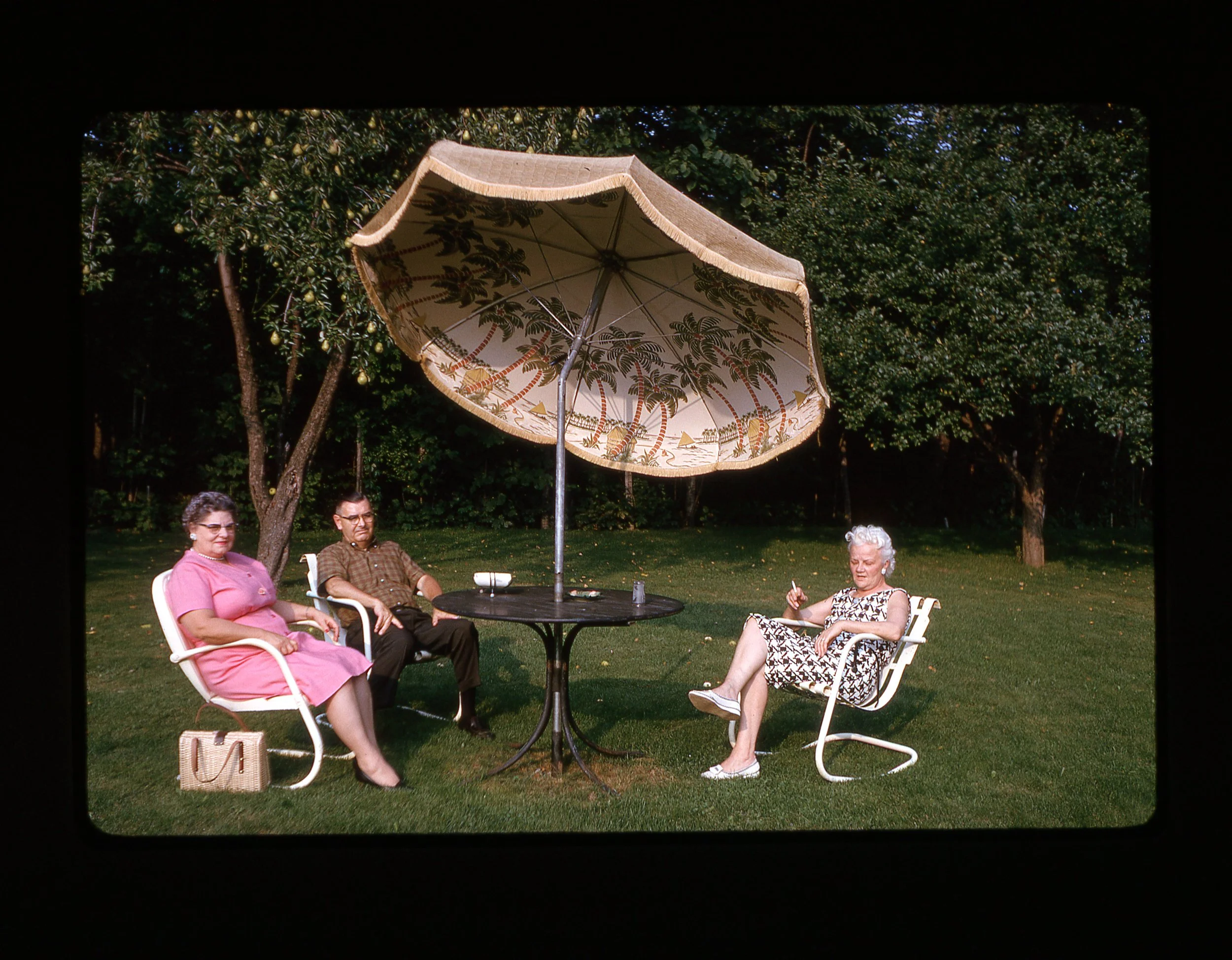 Four people sitting outdoors around a table with a large fabric parasol. The setting appears to be a garden or park with trees and grass.