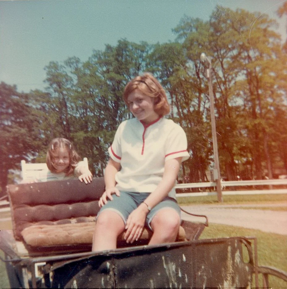 A woman and a young girl sitting on a vintage horse-drawn carriage outdoors, with trees and a road in the background.