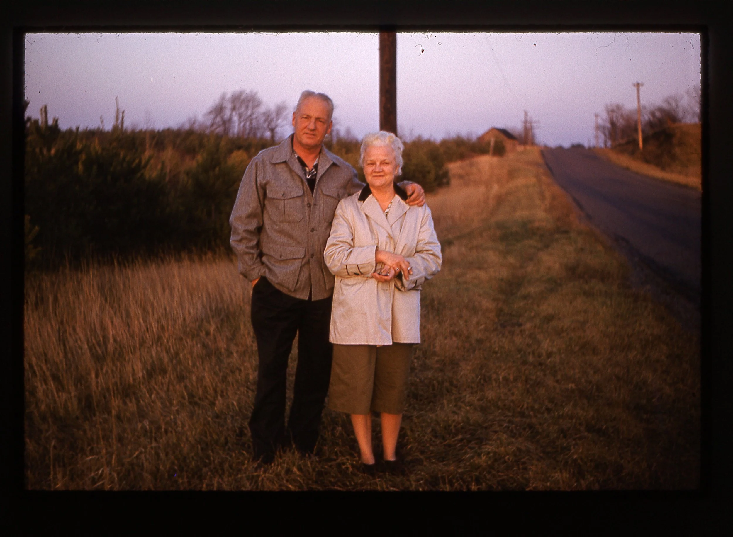 A man and a woman standing close together outdoors on a rural road during sunset. The man has gray hair, wearing a gray jacket, and the woman has white hair, wearing a beige coat.