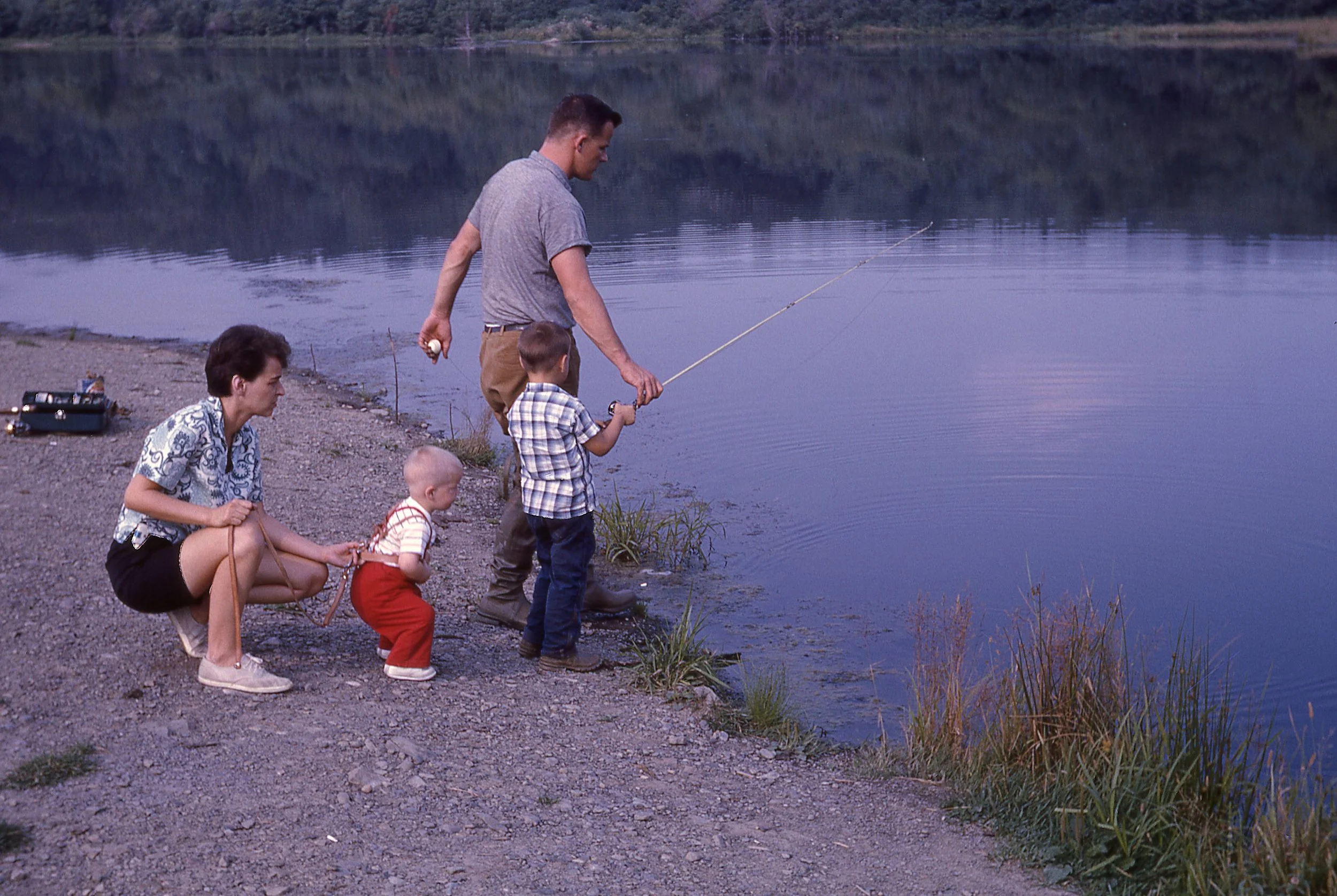 A group of five people, including two children, are fishing by a lake on a rocky shore, with trees and hills in the background during twilight.