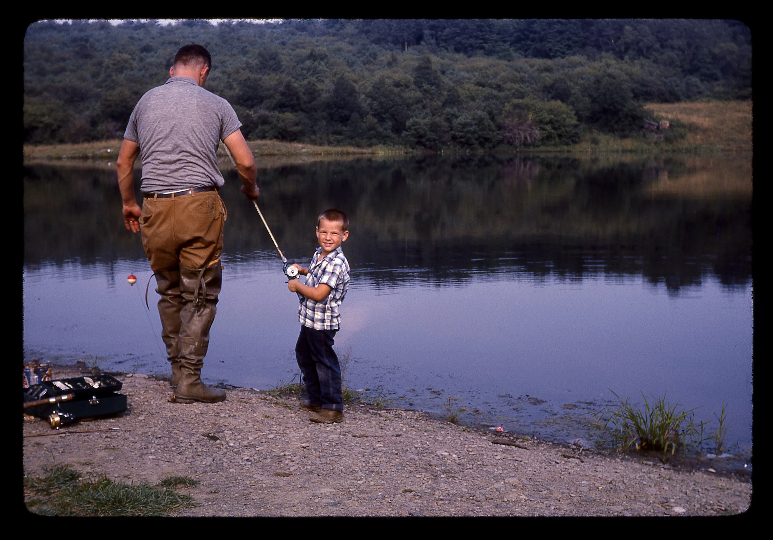 A man and a boy stand by the edge of a lake, with trees and hills in the background, fishing together.