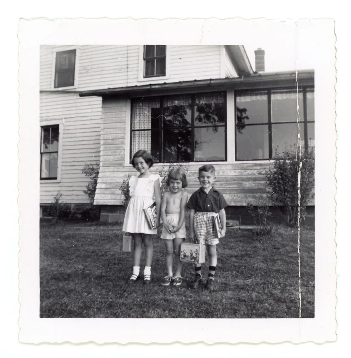 Three children standing on a lawn in front of a house, holding school bags, smiling for the camera in black and white.