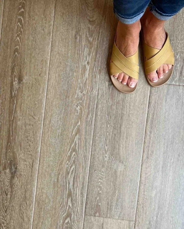 Close-up of a person's feet in yellow sandals and rolled-up jeans standing on wood flooring.