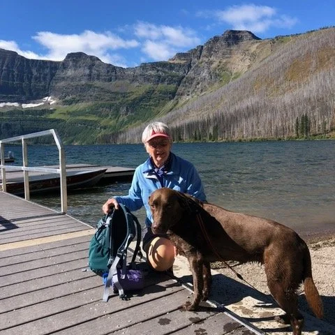 Person and a dark-colored dog resting on a wooden dock by a mountain lake.