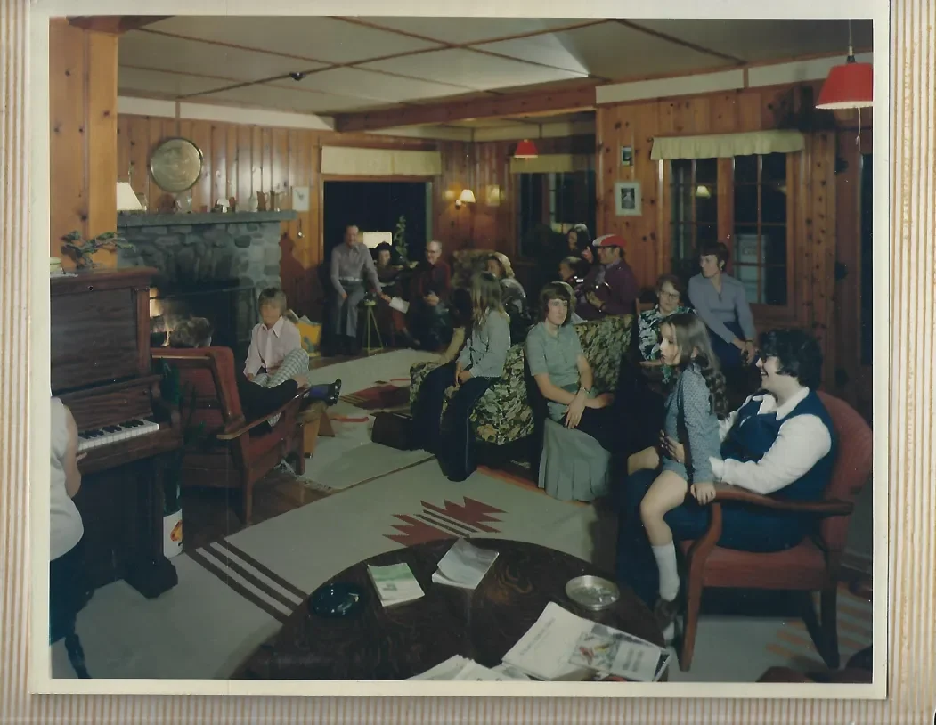 Historic lodge gathering room with wood-paneled walls, a stone fireplace, piano, and guests assembled for an evening of music and socializing.