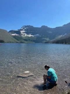 Visitor sitting along the rocky shoreline of a crystal-clear alpine lake with snow-capped mountains in the background at Glacier National Park.