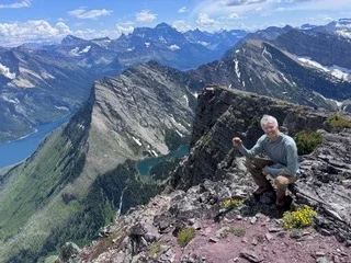 Hiker posing on a rocky mountain summit overlooking dramatic peaks and glacial lakes in Glacier National Park.