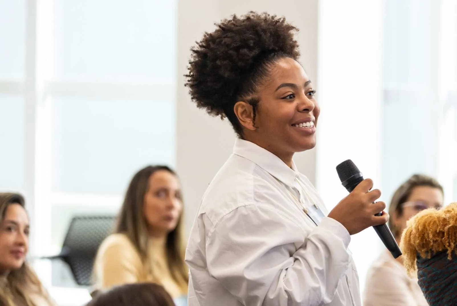 Confident woman stood at smiling holding a microphone whilst audience around here is seated.