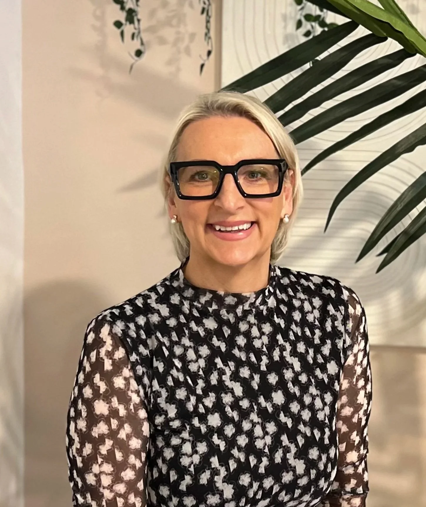 A woman is looking directly at the camera smiling confidently, with black frame glasses, wearing a white and black print dress.