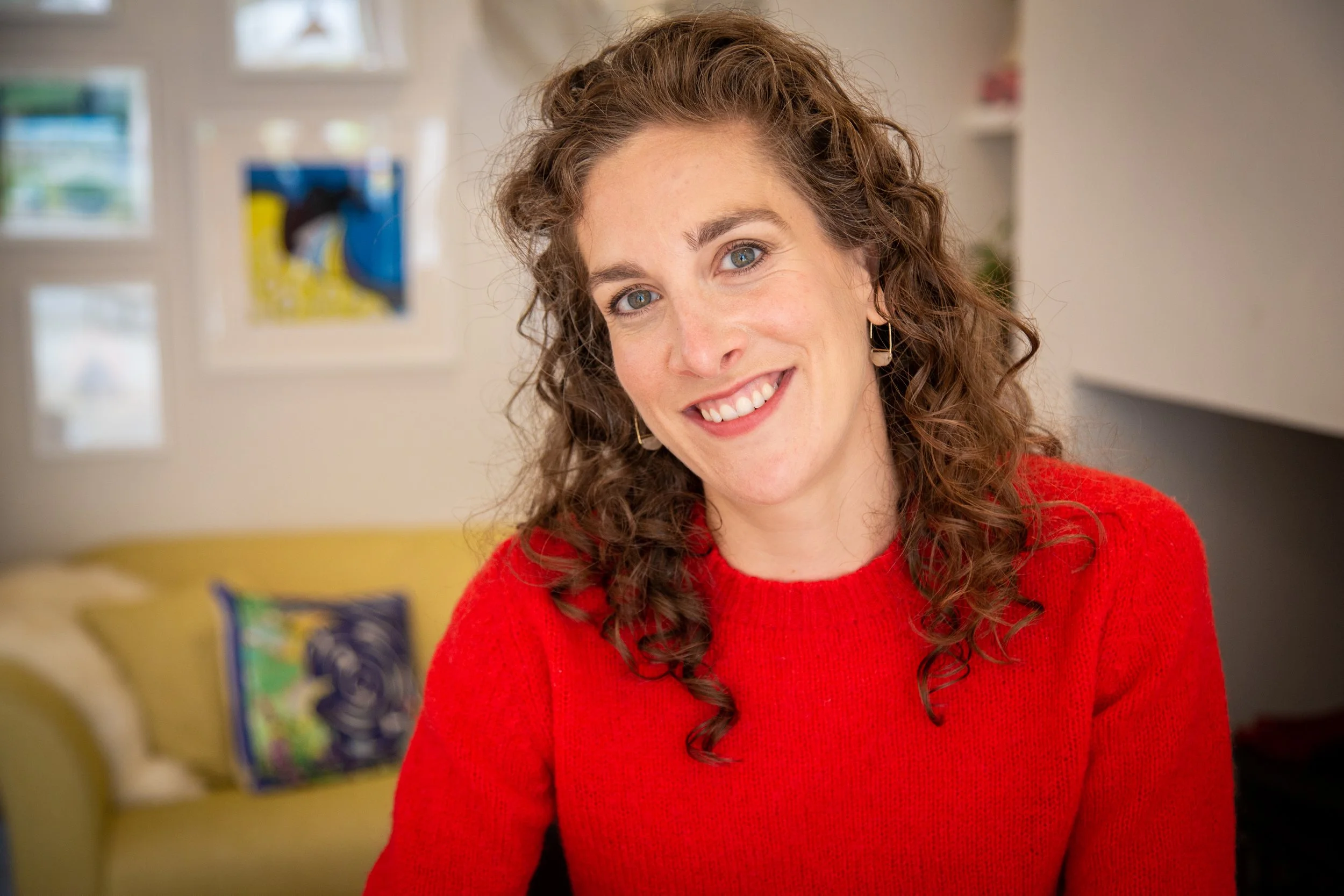 A woman with brown curly hair is staring directly at the camera with a warm smile, she is wearing a bright red jumper.