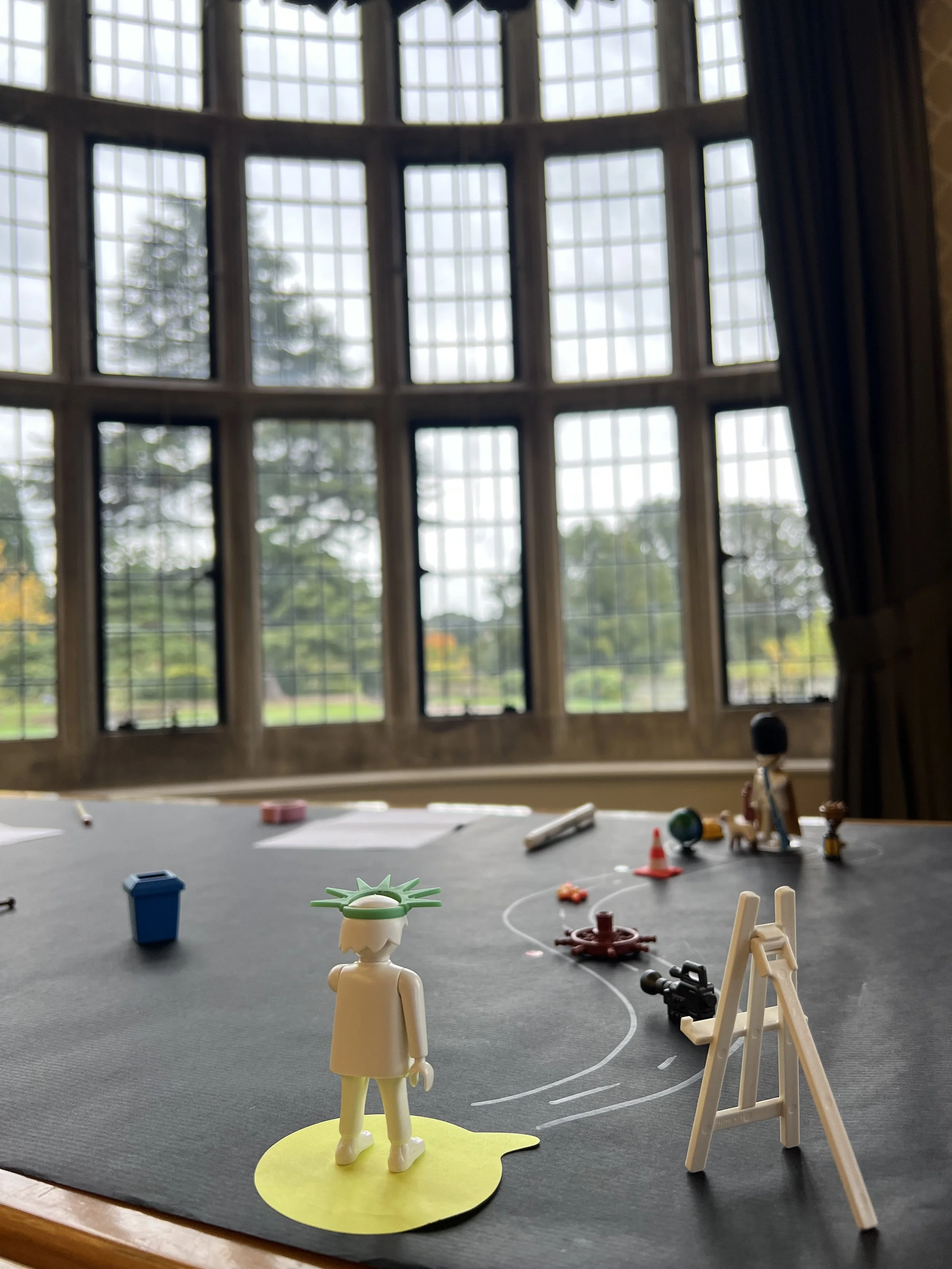 A scene is laid out on a black table, with a chalk outline of a roadmap. Playmobil characters and pieces are along the path. In the background is a grand window overlooking autumnal trees.