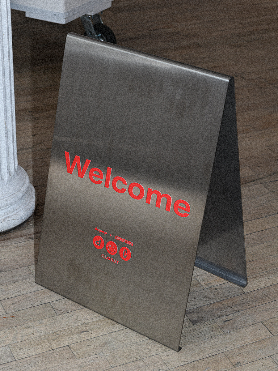 A metal A-frame sign with red text that says "Welcome" and icons for DEPOP and COSMOPOLITAN brands on a wooden floor inside a building.