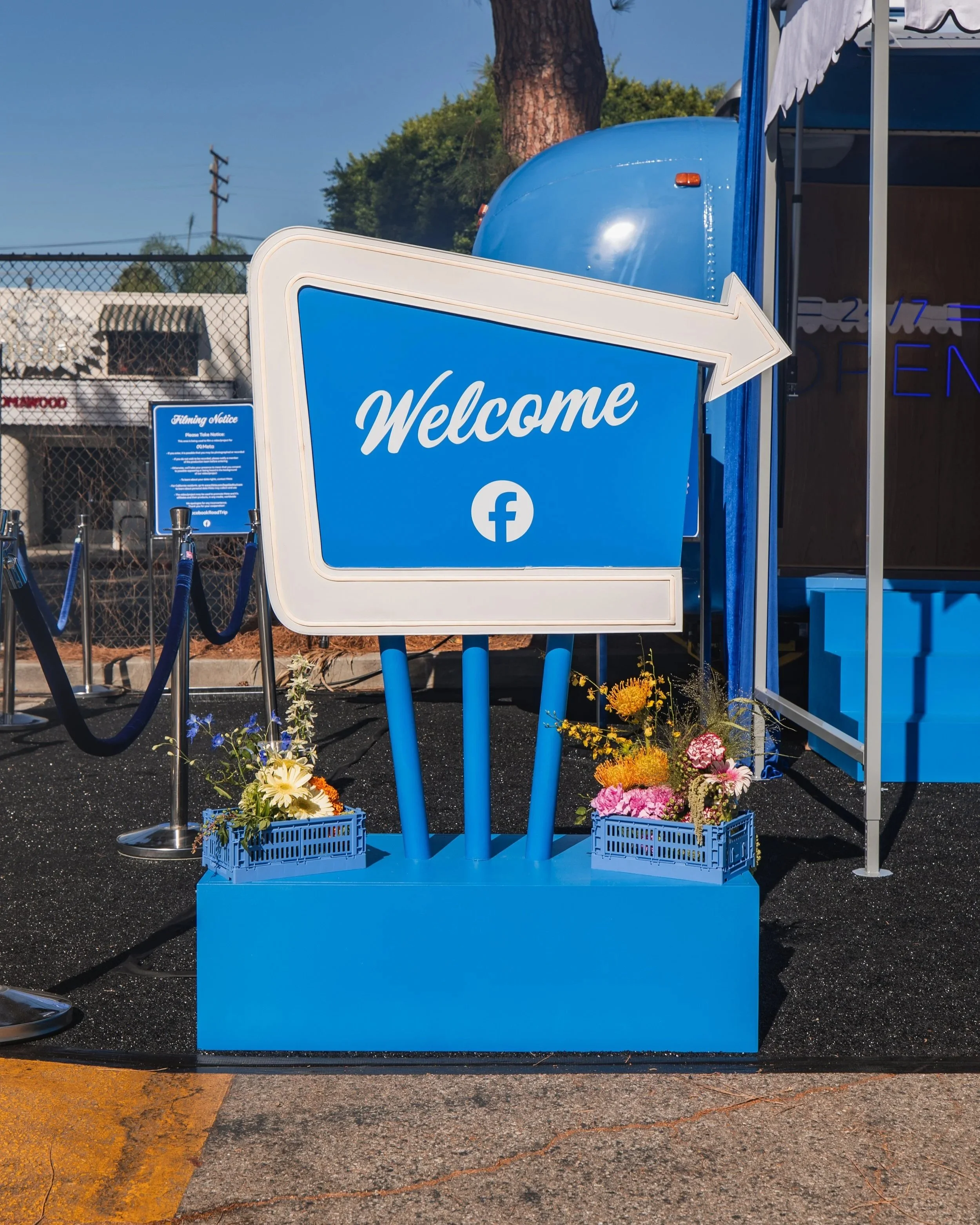 A blue welcome sign with an arrow pointing to the right, situated on a sidewalk with flowers in blue baskets below. Part of a blue trailer with a white canopy is visible to the right. In the background, there is a chain-link fence and signs, with trees and utility poles behind.