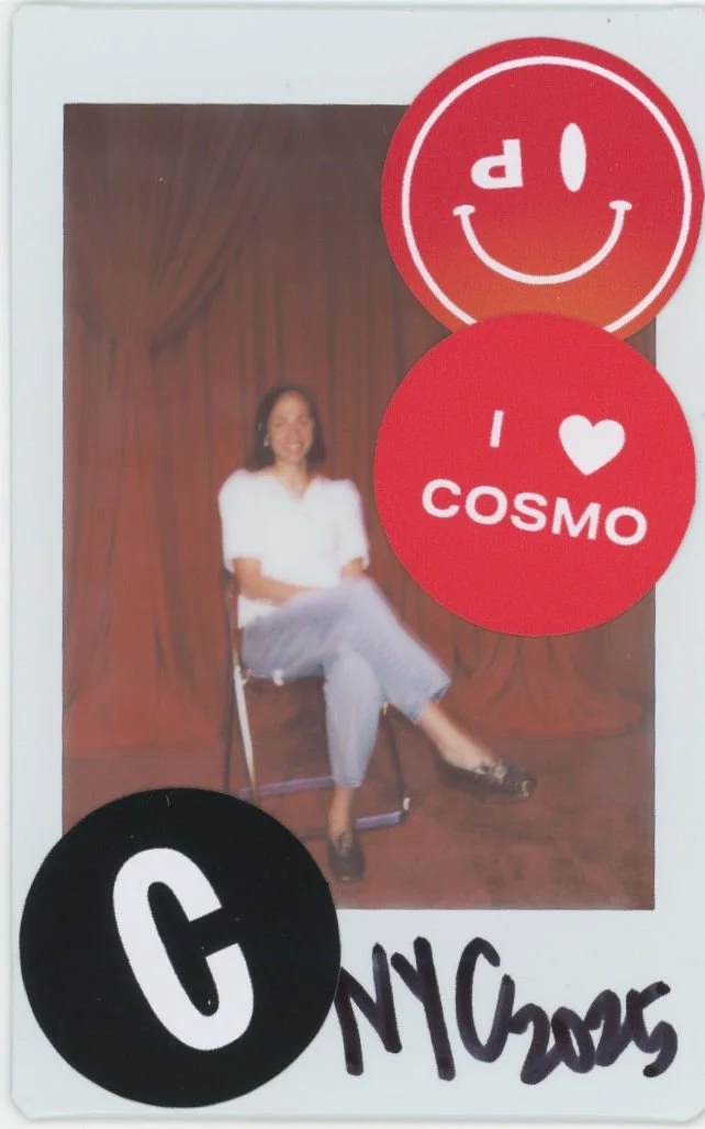 A polaroid image of a woman sitting on a chair in front of a brown curtain. There are round stickers on the photo with DEPOP and COSMOPOLITAN brand logos and smiling faces. The bottom part of the photo has handwritten text that says 'NYC 2023.'