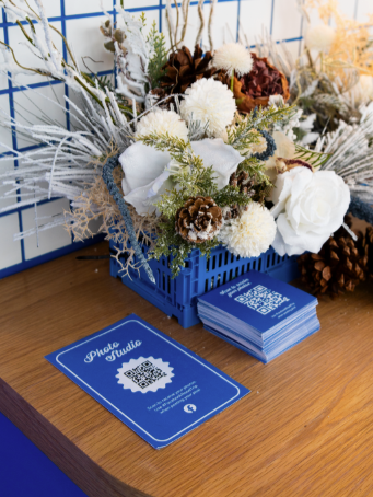 A blue basket holding artificial white flowers and brown pinecones, placed beside a stack of blue business cards with a white QR code and text, on a wooden surface.
