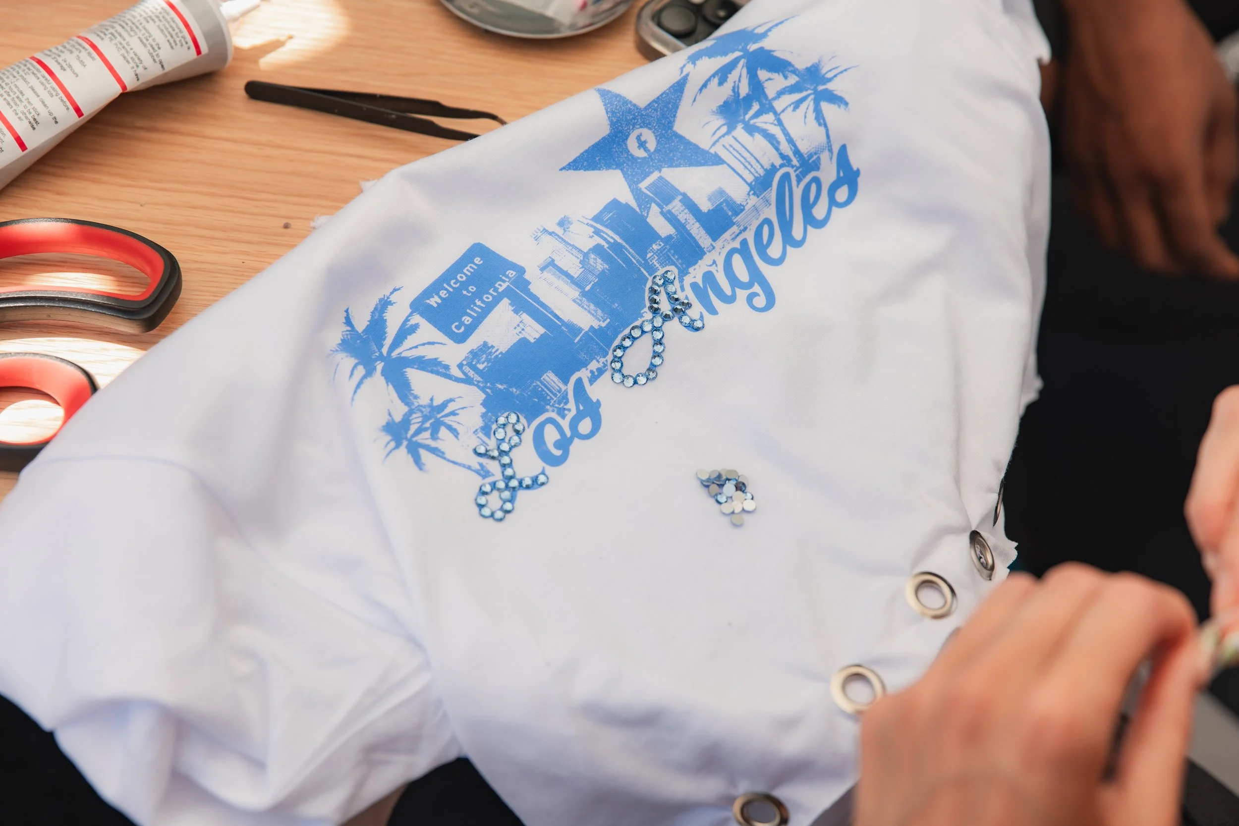 A white T-shirt with a blue graphic design of Los Angeles landmarks and palm trees, with the words 'Welcome to California' and 'Los Angeles' written on it. The T-shirt is placed on a wooden surface alongside scissors, a container, and some rhinestones.