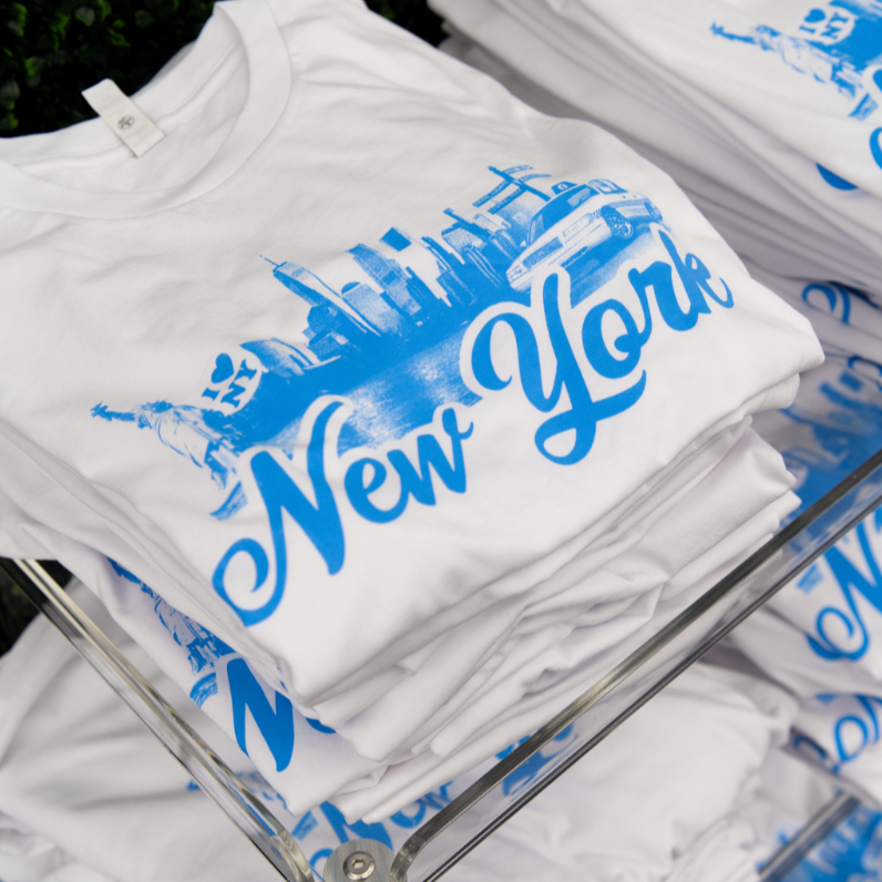 White T-shirt with blue graphic of New York City skyline, Statue of Liberty, police car, and the words 'New York' in large cursive font.
