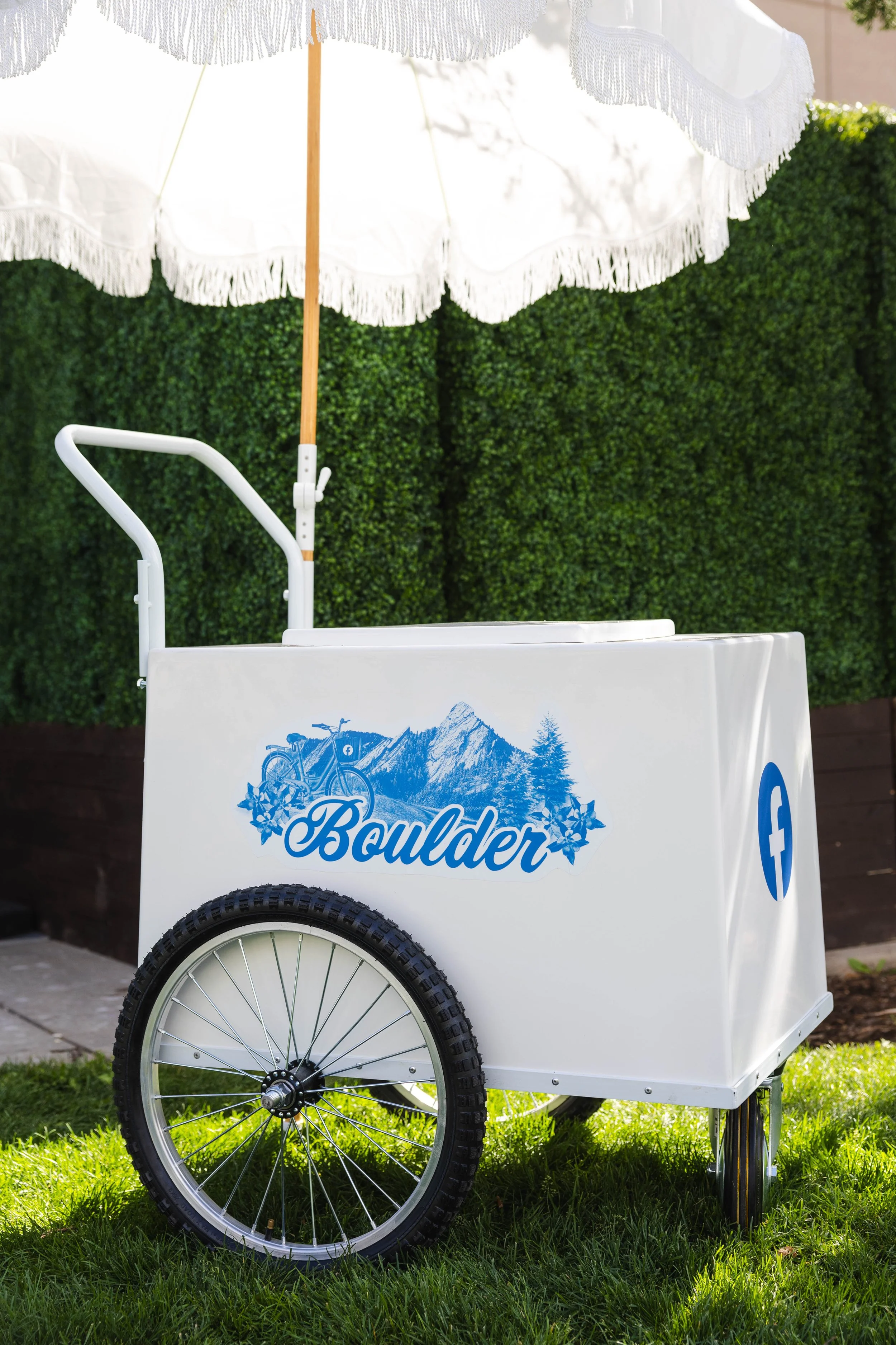 A white Boulder ice cream cart with two large wheels, underneath a white fringed parasol, set against a green hedge, with a mountain and trees graphic on the side of the cart.