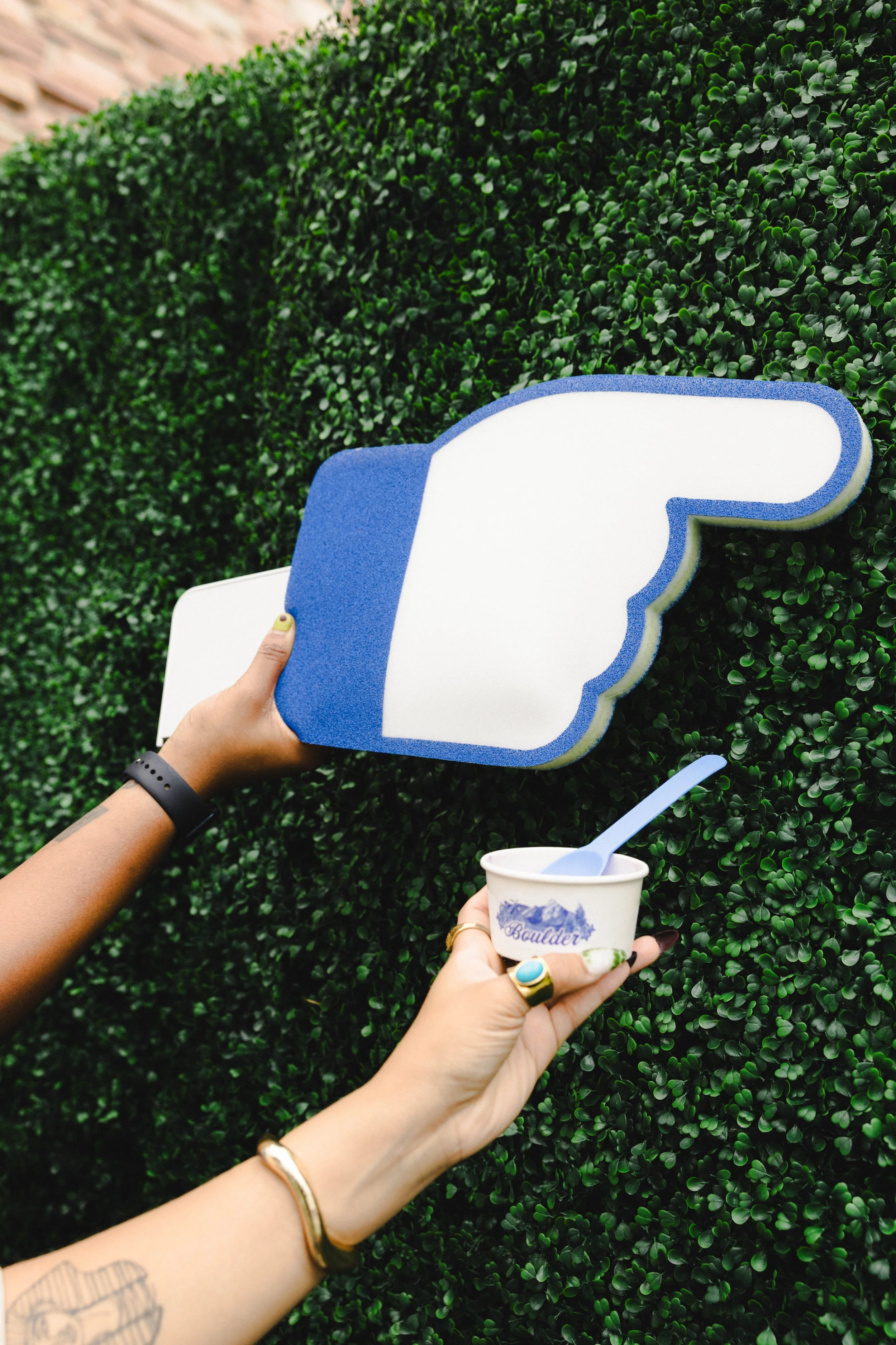 Hands holding a foam finger shaped like a thumbs-up sign and a cup of ice cream with a spoon in front of a green hedge.