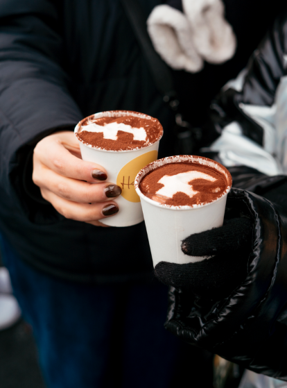 Two people holding cups of hot chocolate topped with whipped cream in Facebook logo shape and cocoa powder.
