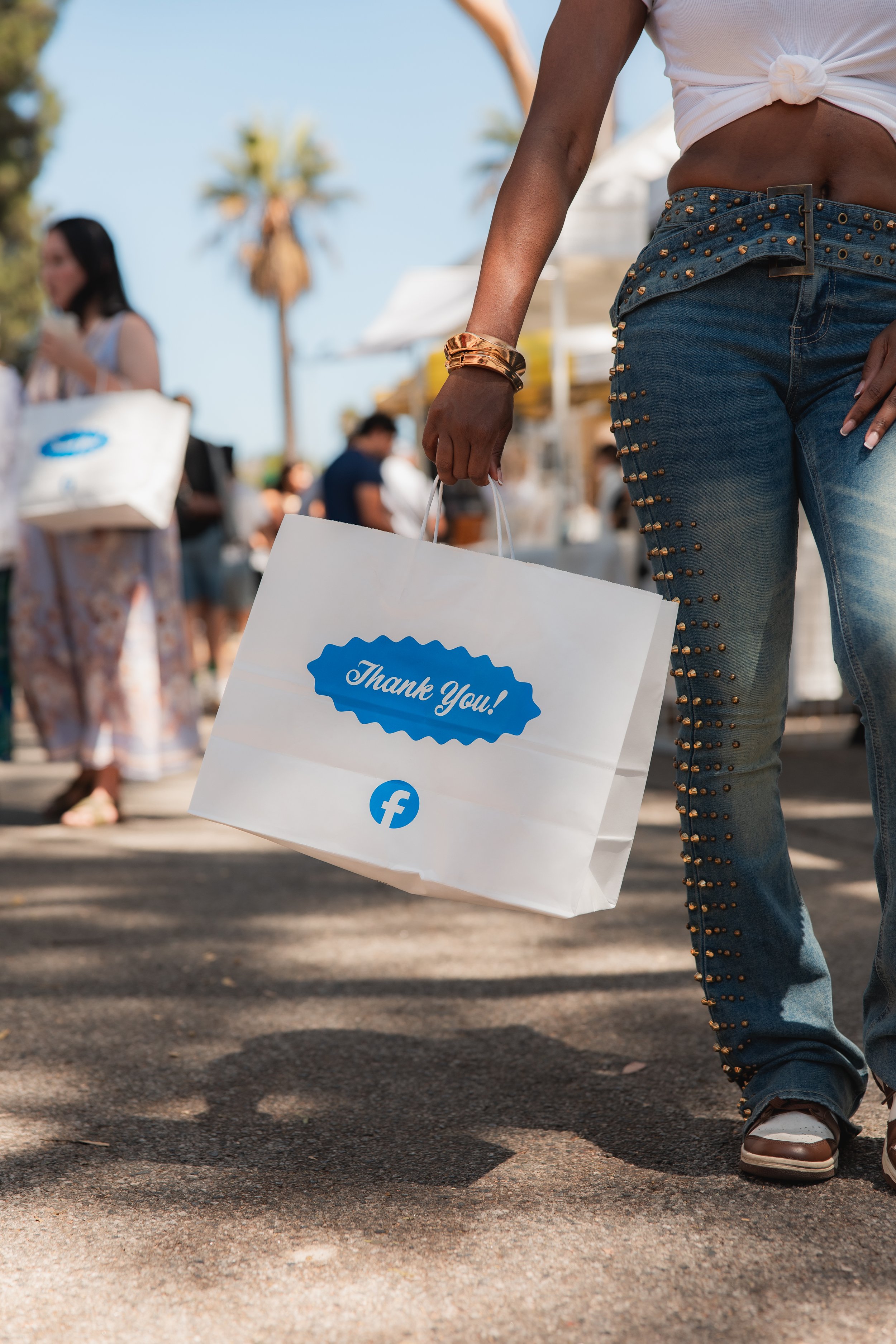 A person is walking outdoors carrying a shopping bag with a "Thank You!" message and a Facebook logo. They are wearing jeans with studs along the side, a tied white shirt, and brown shoes. In the background, there are people and palm trees.