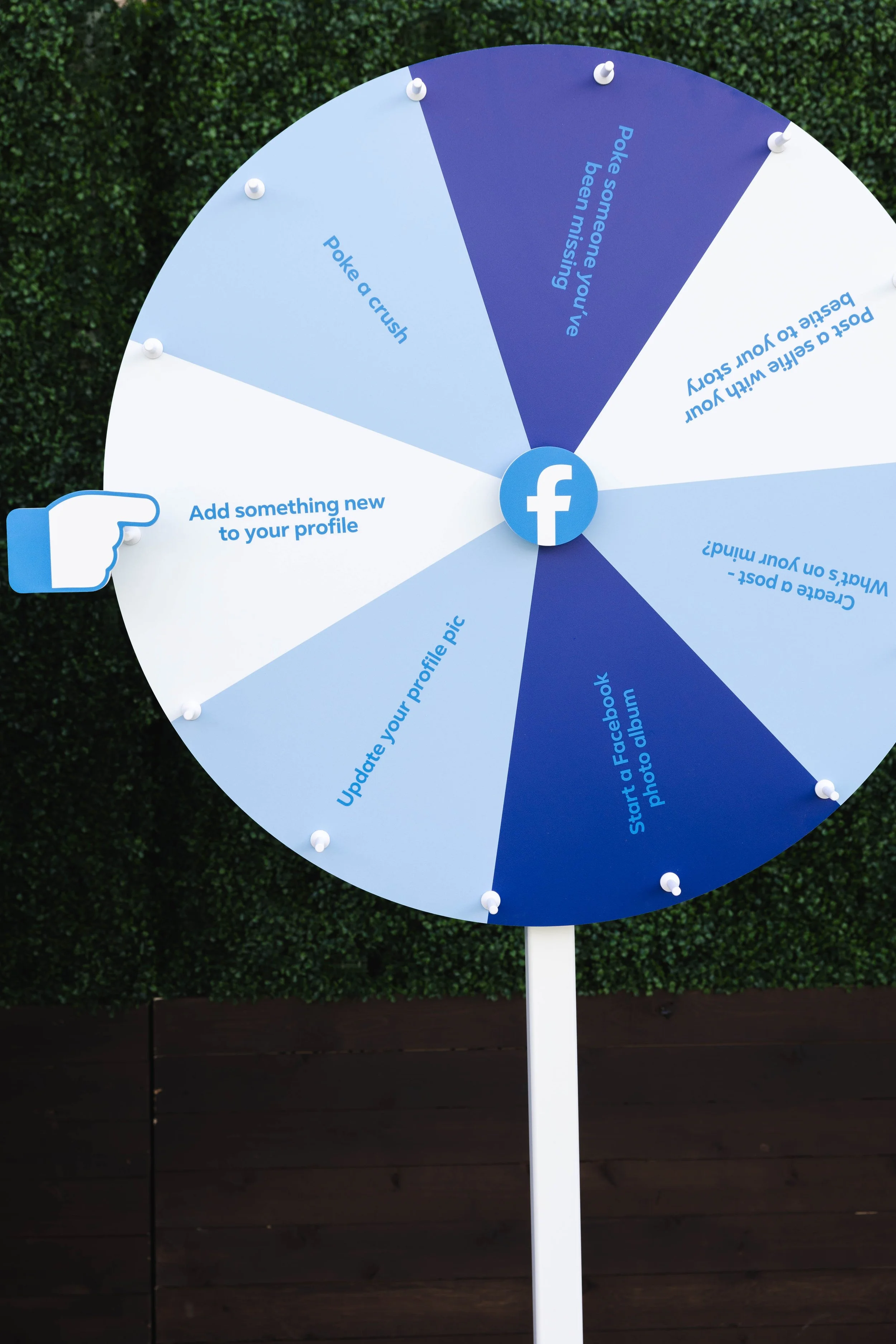 A circular Facebook-themed wheel with various suggestions about using Facebook, including to add something new to your profile, update your profile picture, start a Facebook photo album, post a crush, poke someone you’ve been missing, tell a Facebook story, or describe your week in an email. The wheel is mounted on a white pole in front of a green hedge.