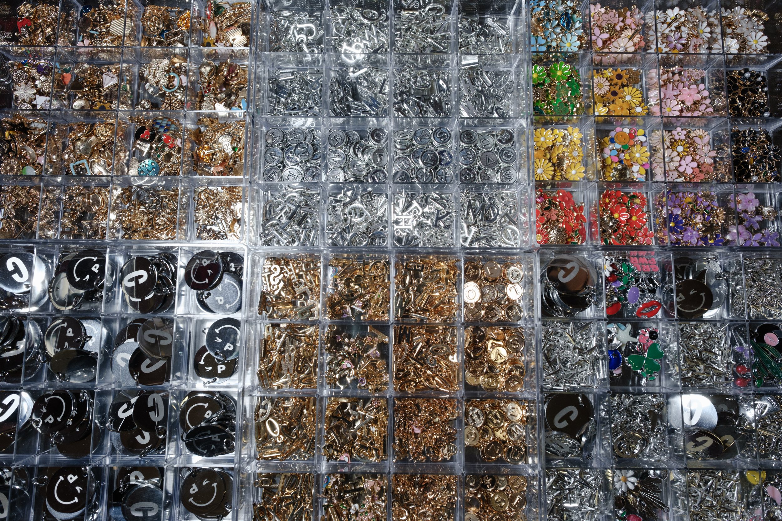 A plastic storage box with multiple compartments filled with colorful metal charms and jewelry-making supplies, including lettered alphabet beads, flower-shaped charms, and various other decorative pieces.