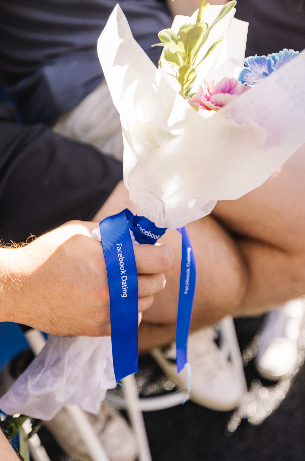 Person holding a wedding bouquet wrapped in white paper and tied with a blue ribbon that says 'Facebook Dating'.