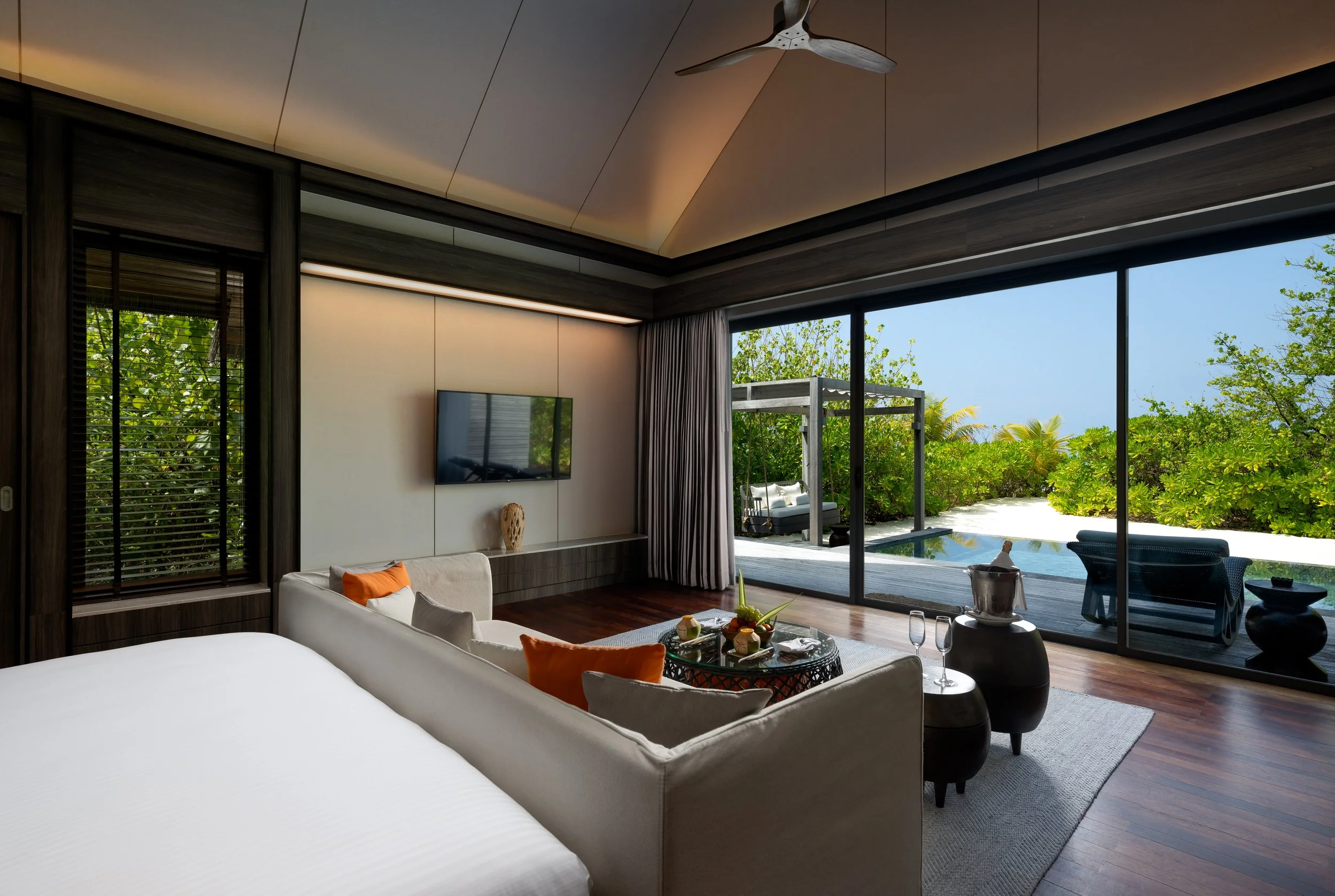 Modern living room with large glass sliding doors opening to a lush garden and outdoor seating area, featuring a contemporary ceiling fan, wall-mounted TV, and minimalist décor.