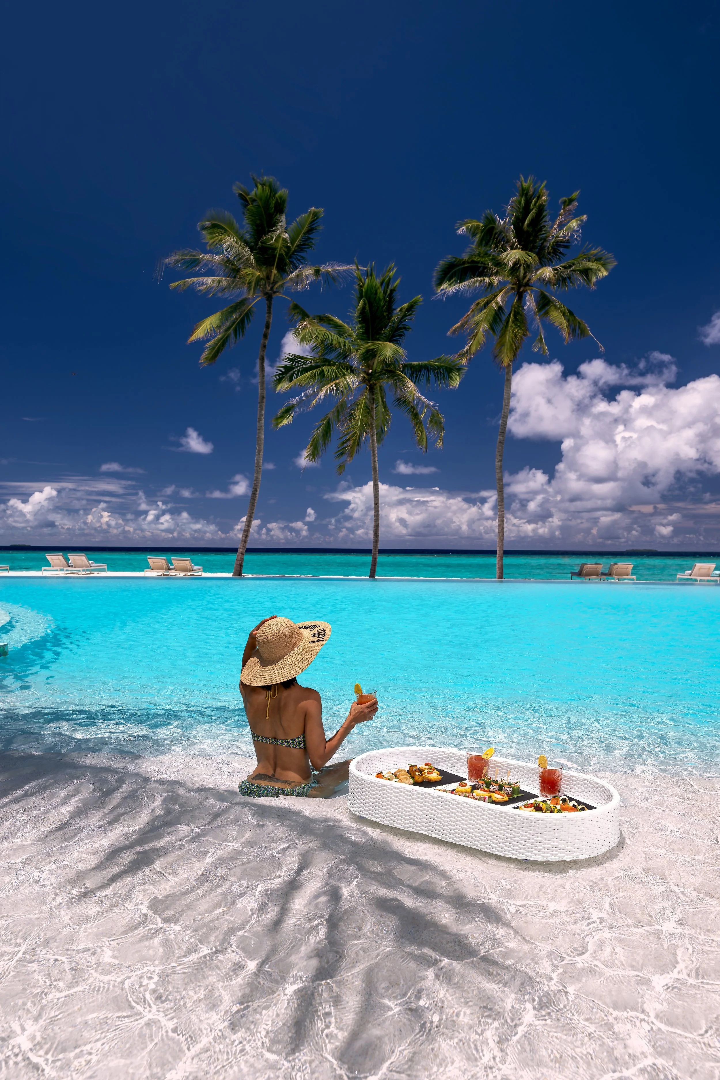 A woman sitting on the beach by the ocean, wearing a sun hat and bikini, enjoying a meal with drinks and a tray of food on the sand, with palm trees and lounge chairs in the background under a partly cloudy sky.