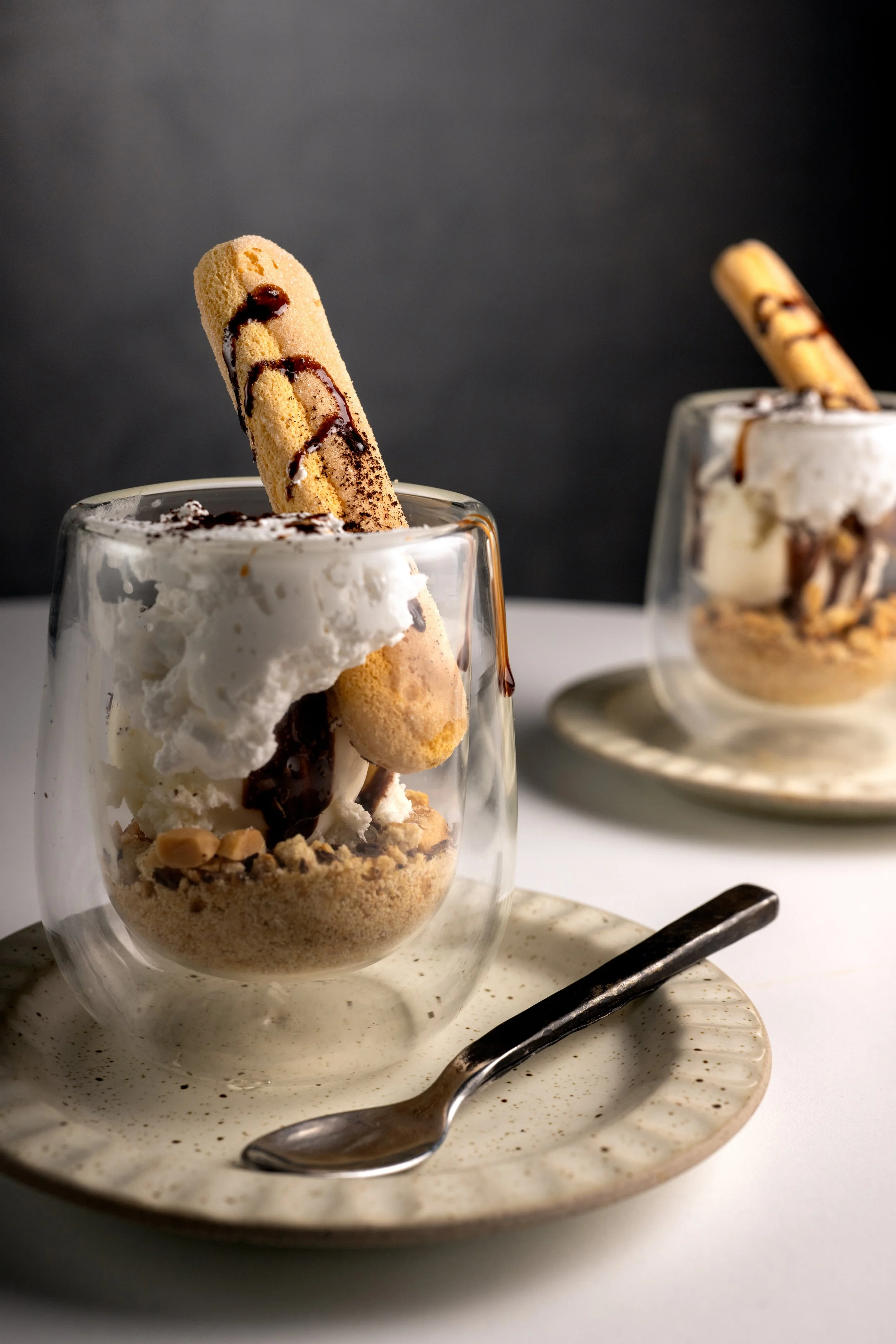 A glass of ice cream topped with whipped cream, chocolate syrup, and a biscotti, served on a speckled ceramic plate with a spoon.