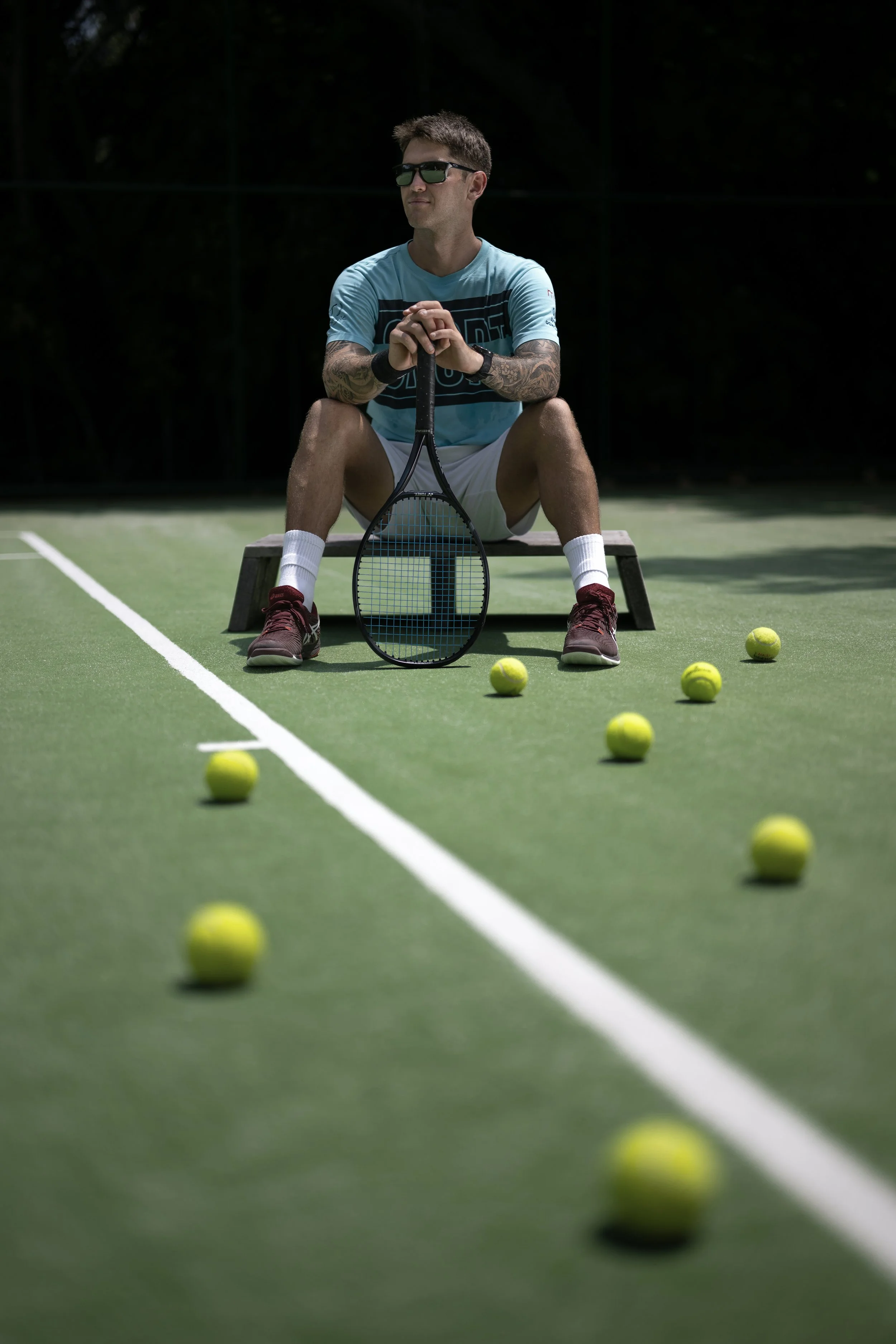 A man sitting on a bench on a tennis court, surrounded by tennis balls, holding a tennis racket, wearing sunglasses, a blue shirt, white shorts, and red shoes.
