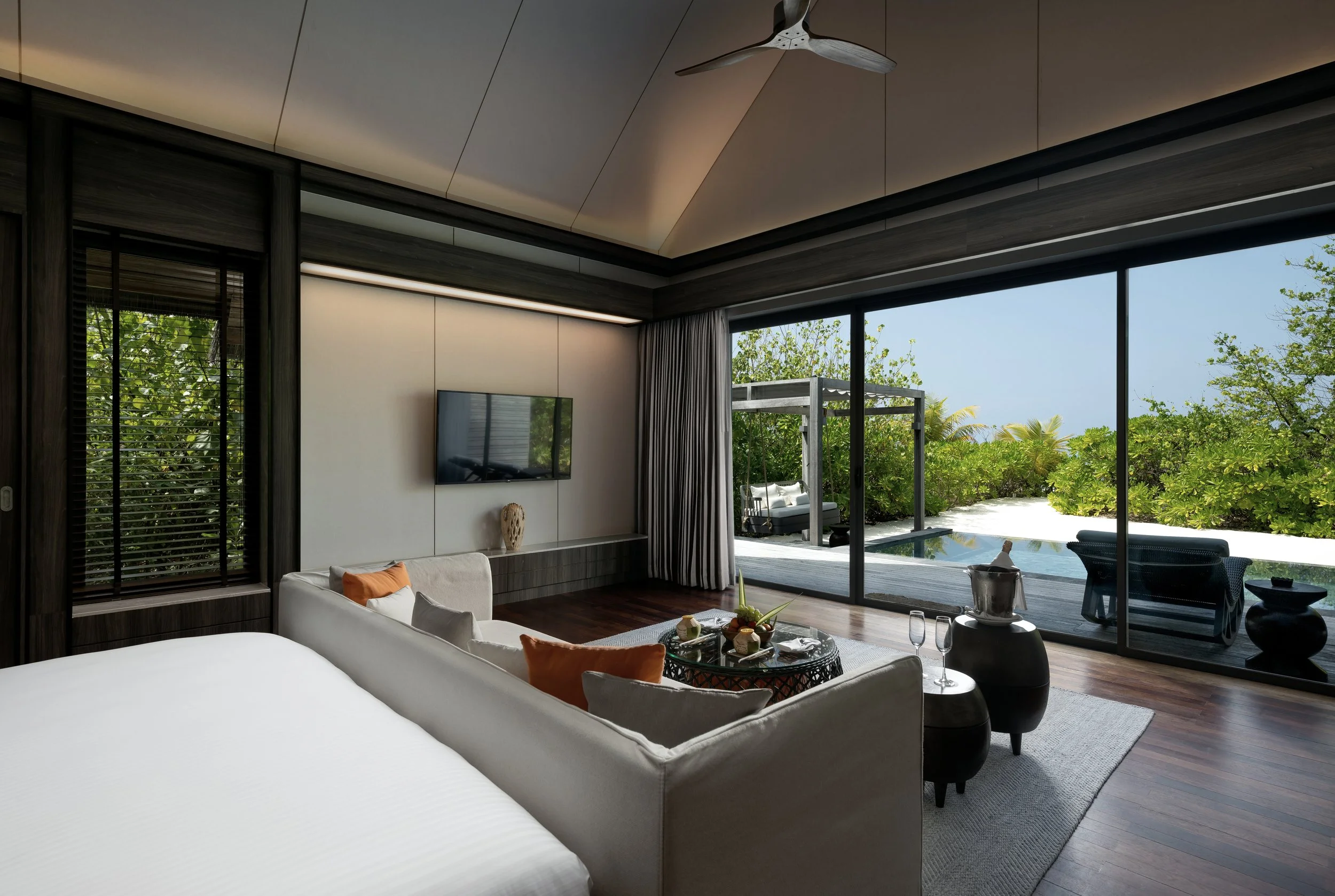 Modern living room with large glass sliding doors leading to an outdoor patio with greenery and pool, featuring white sofas with orange pillows, a wall-mounted TV, wooden walls, and a ceiling fan.