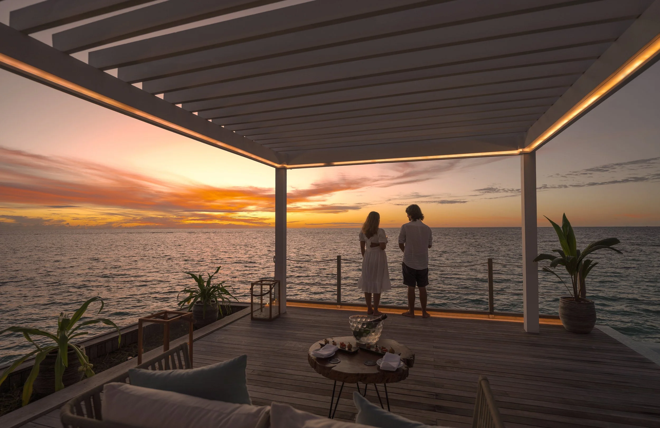 A man and woman standing on a balcony overlooking the ocean during sunset, with a modern roof structure and potted plants in the foreground.
