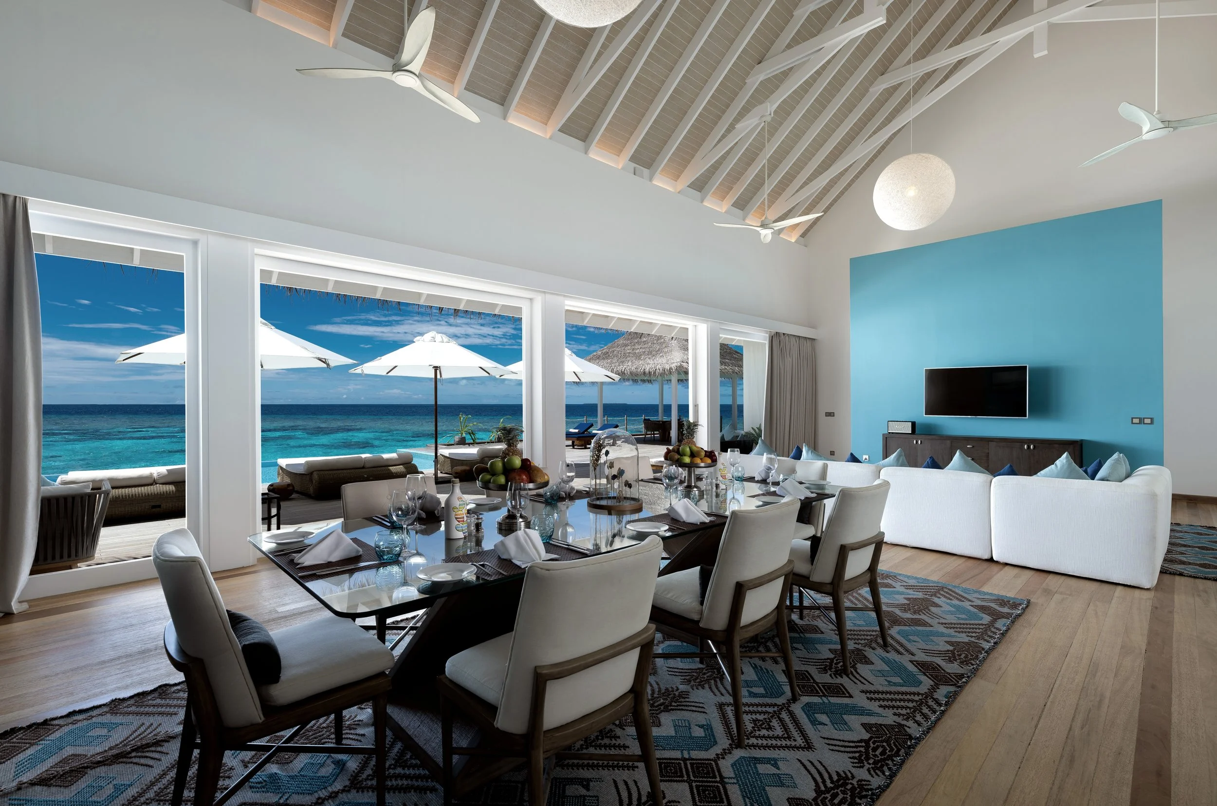 An interior living and dining room with large sliding glass doors opening to a beach view with umbrellas and blue ocean. The room has white walls, wooden flooring, a patterned area rug, a dining table with chairs, a sofa, a television, modern ceiling