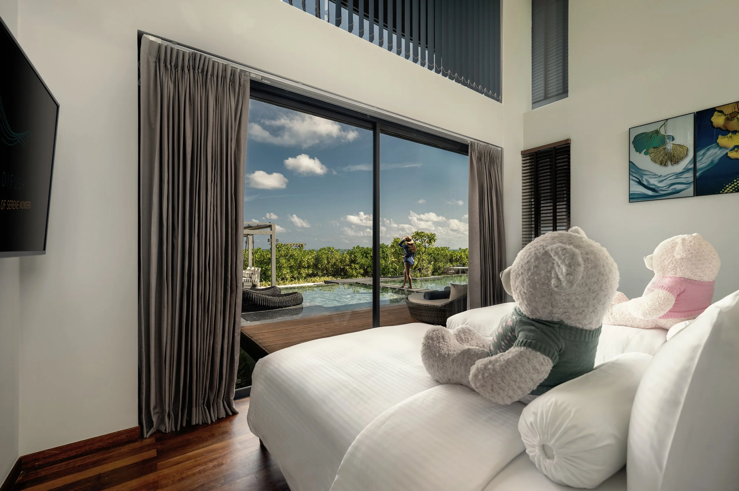 A bedroom with two teddy bears sitting on a bed, facing a large glass door that opens to a patio with an outdoor seating area and a swimming pool. The patio overlooks a lush green landscape under a partly cloudy sky.