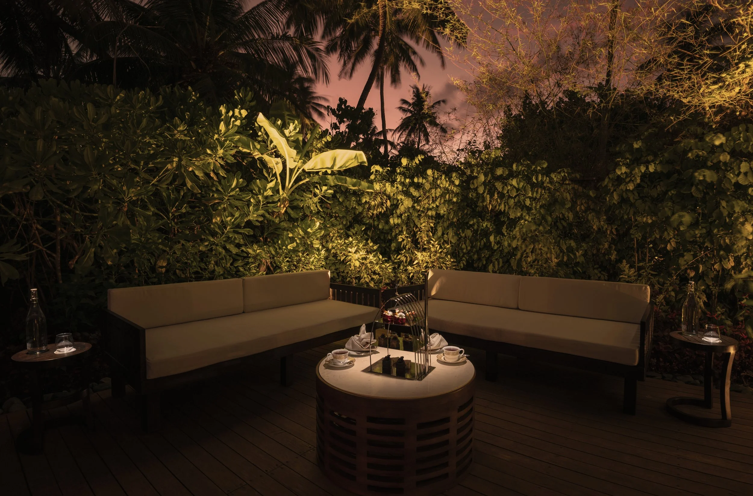 Outdoor patio area at night with cushioned seating, small tables with drinks, and lush green plants and trees illuminated by soft lighting.