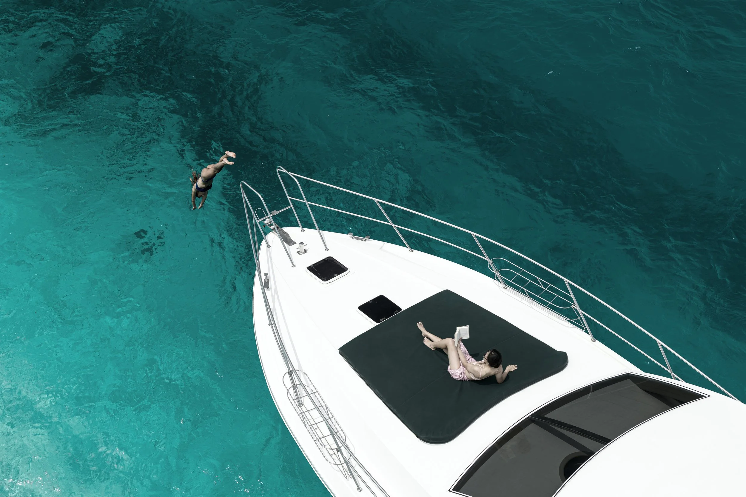 A woman wearing a pink dress lies on her back on a black cushioned lounge on the deck of a white yacht, relaxing on a sunny day with turquoise water.