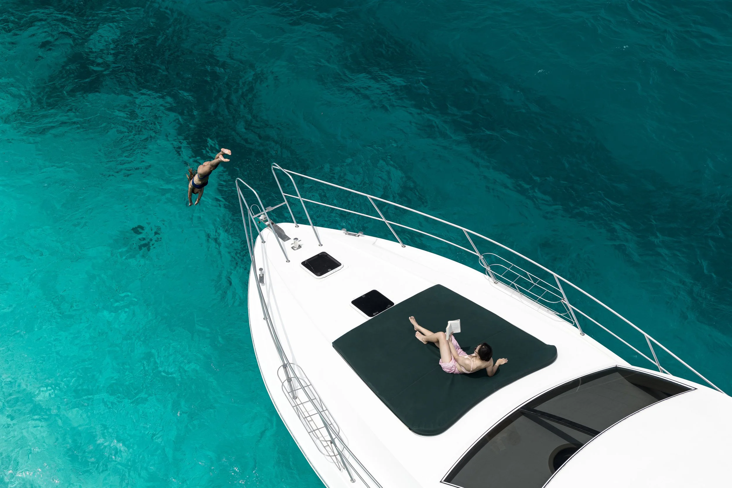Aerial view of a woman diving off a sailboat into turquoise water with another woman lying on a sunbathing mat on the boat.