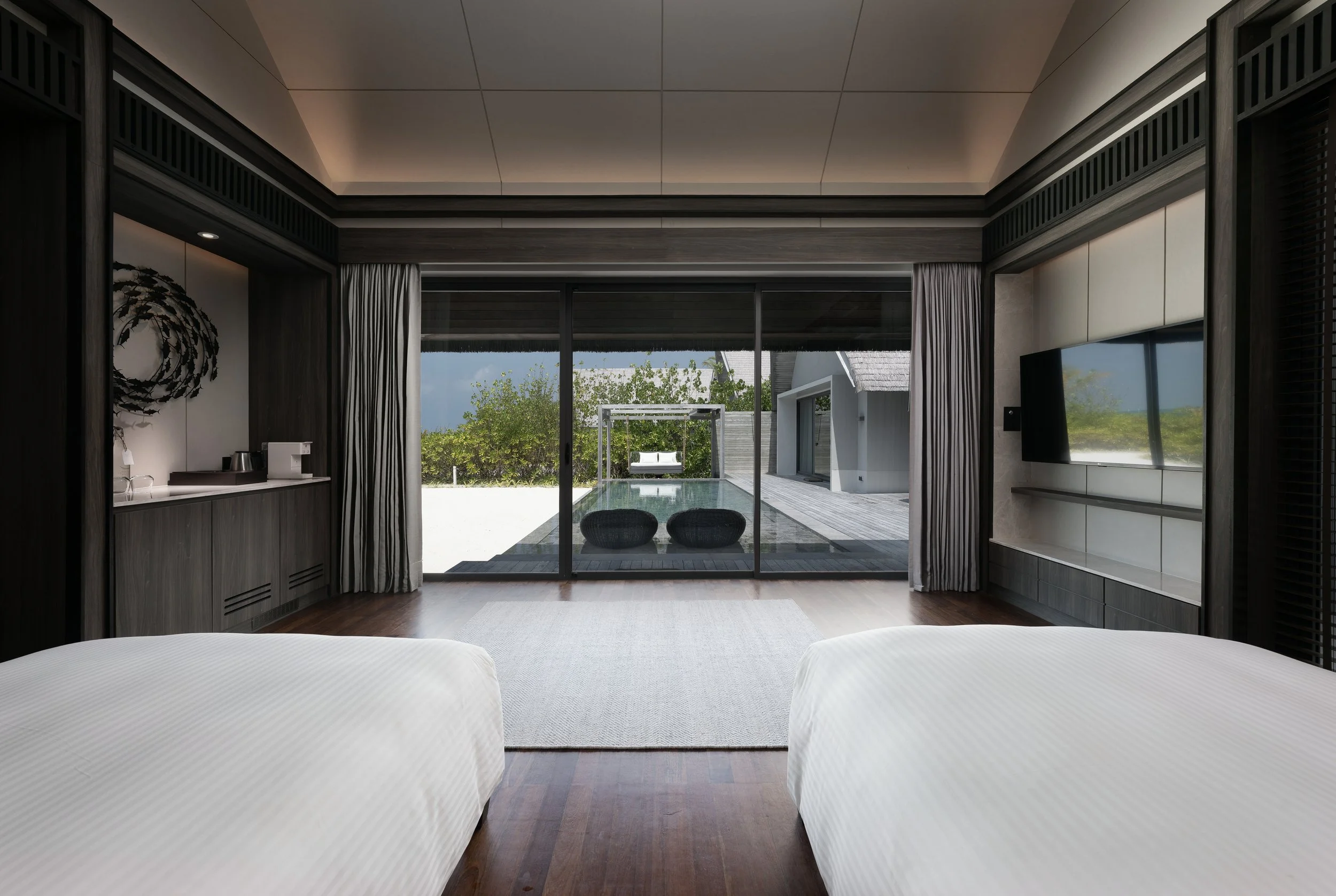 Modern bedroom with two beds facing a glass door leading to an outdoor patio with seating, overlooking a green landscape.