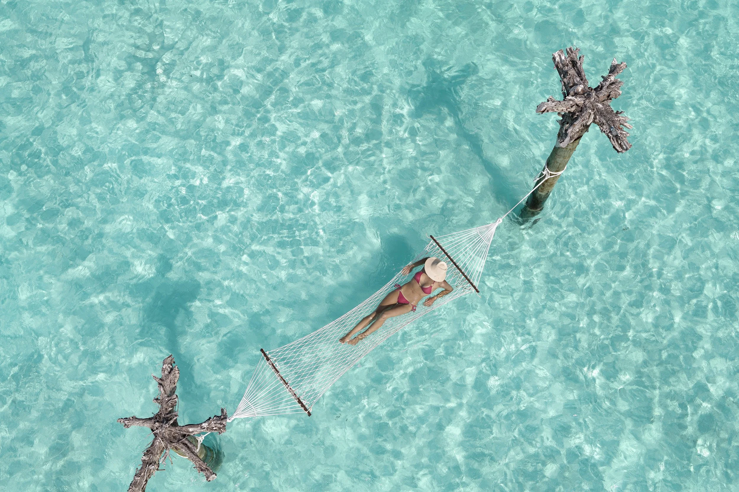 A woman relaxing on a hammock strung between two weathered wooden posts in turquoise water.