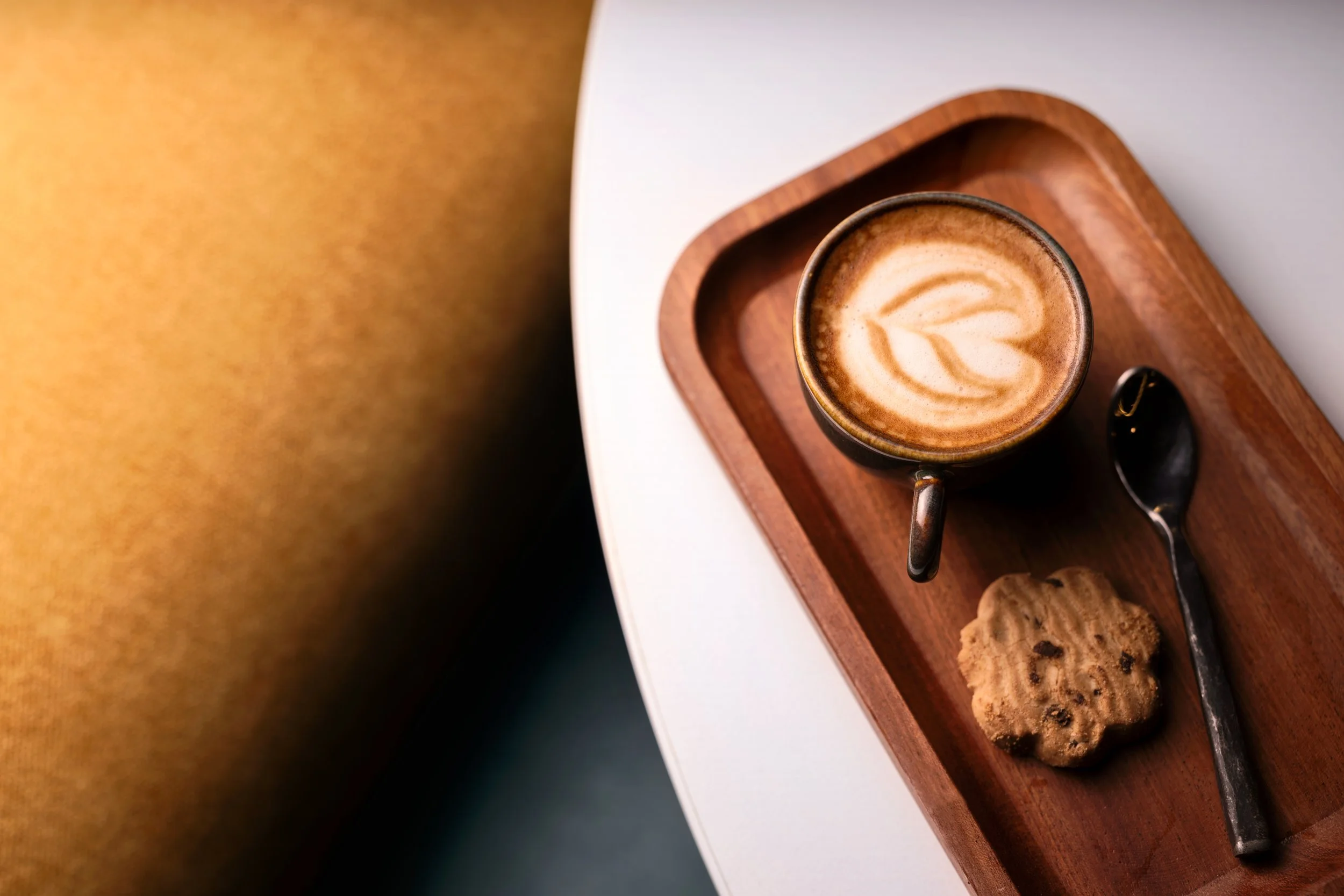 A cup of coffee with latte art on a wooden tray, along with a chocolate chip cookie and a spoon, on a white surface.