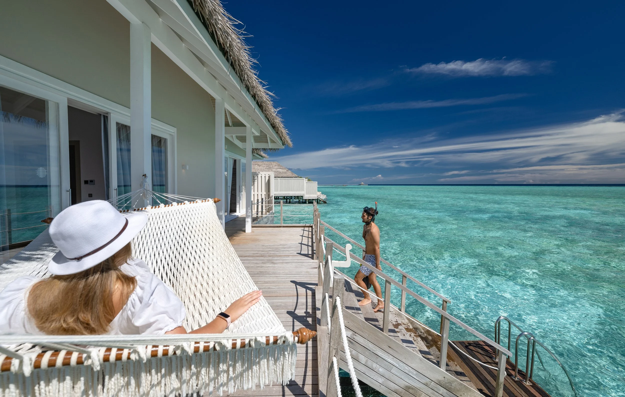 A woman with long hair lying on a white hammock on a wooden deck outside a beachside villa, wearing a wide-brimmed hat. A man wearing swim shorts and snorkel gear stands by the edge of turquoise ocean water, with a distant overwater bungalow and blue