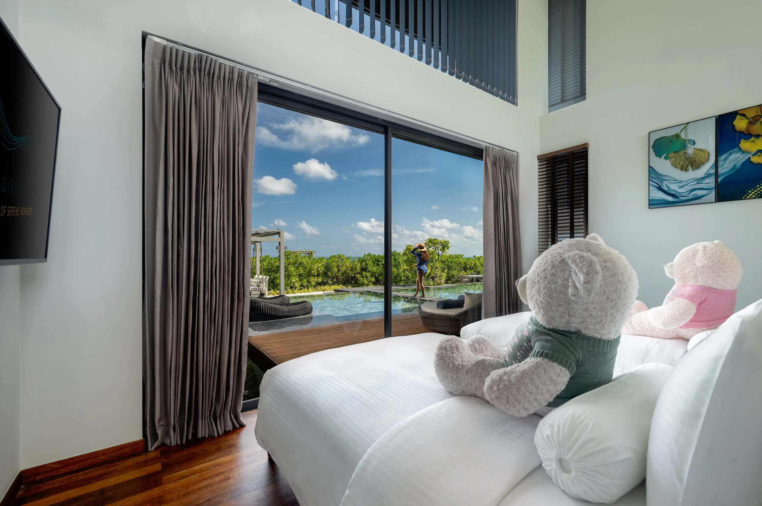 A bedroom with a large window showing a sunny outdoor pool area with green bushes, a person standing outside, and blue sky with clouds. Inside, there are two teddy bears on the bed, one with a pink outfit and the other with a gray and white outfit. T